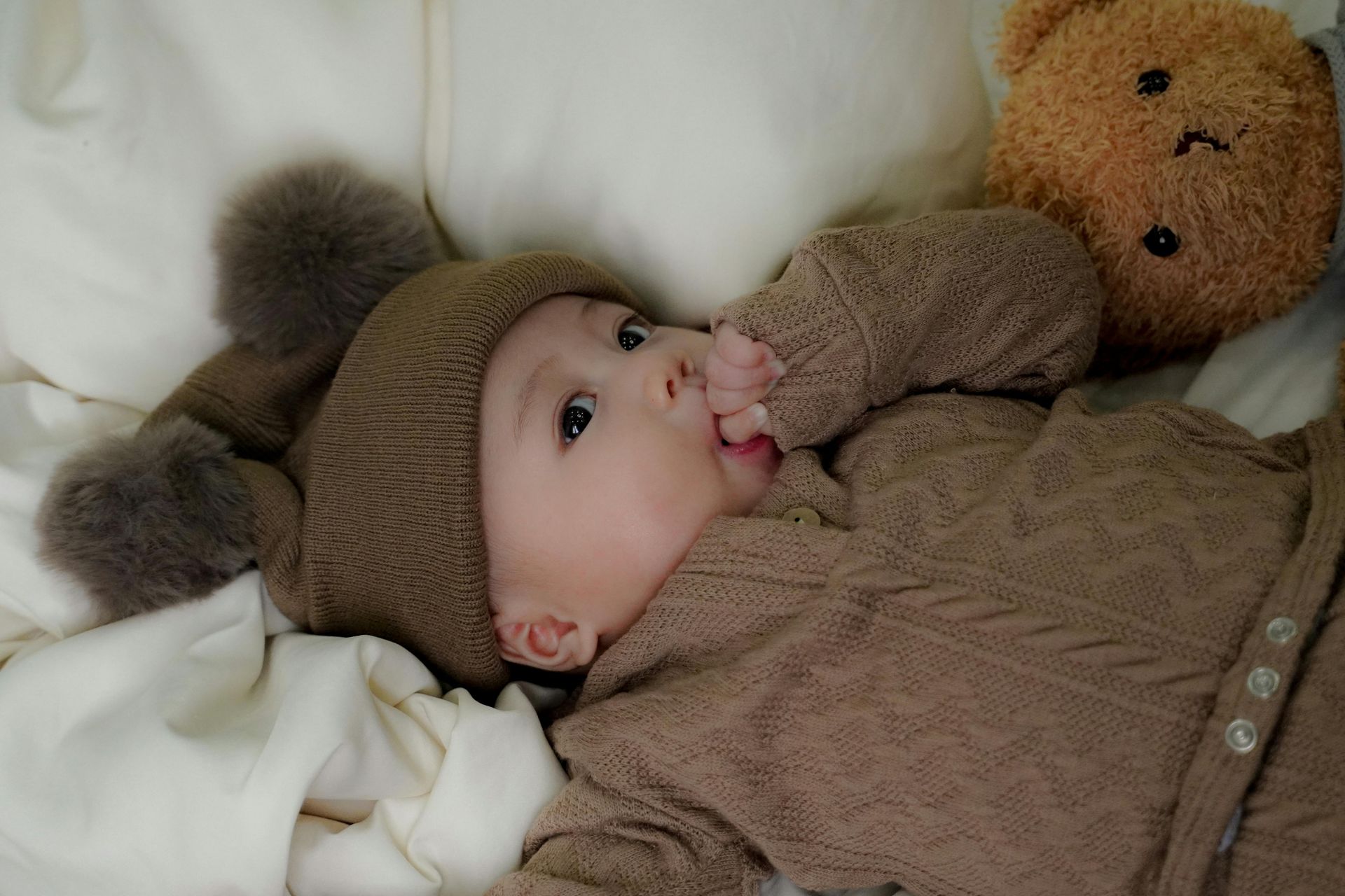 A baby wearing a brown knit hat with pom-poms and a matching sweater lies on a white blanket next to a brown teddy bear.