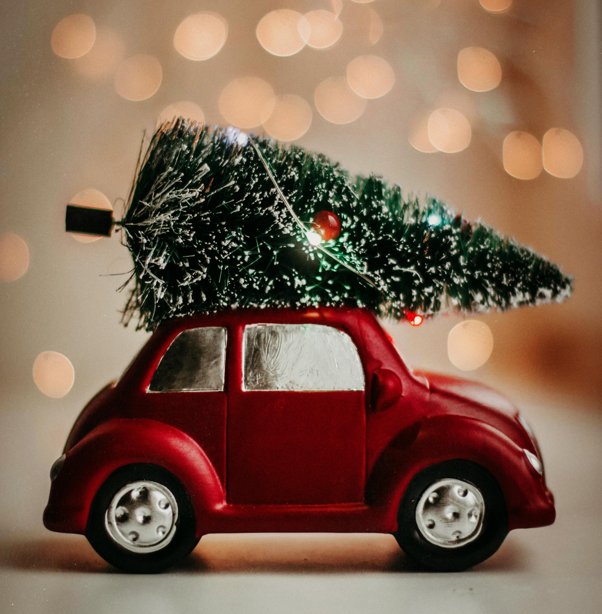 A miniature red toy car carrying a small decorated Christmas tree on its roof against a background of soft bokeh lights.