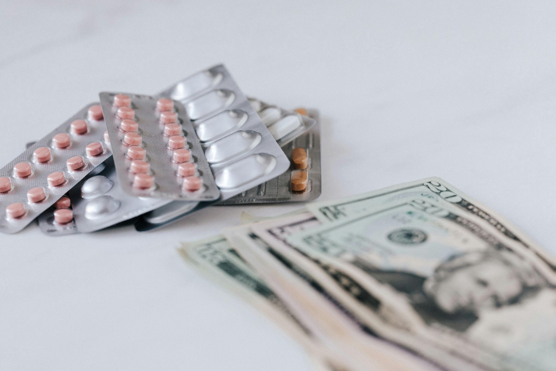 Multiple blister packs of pink and brown medication pills resting next to a stack of US dollar bills on a white surface.