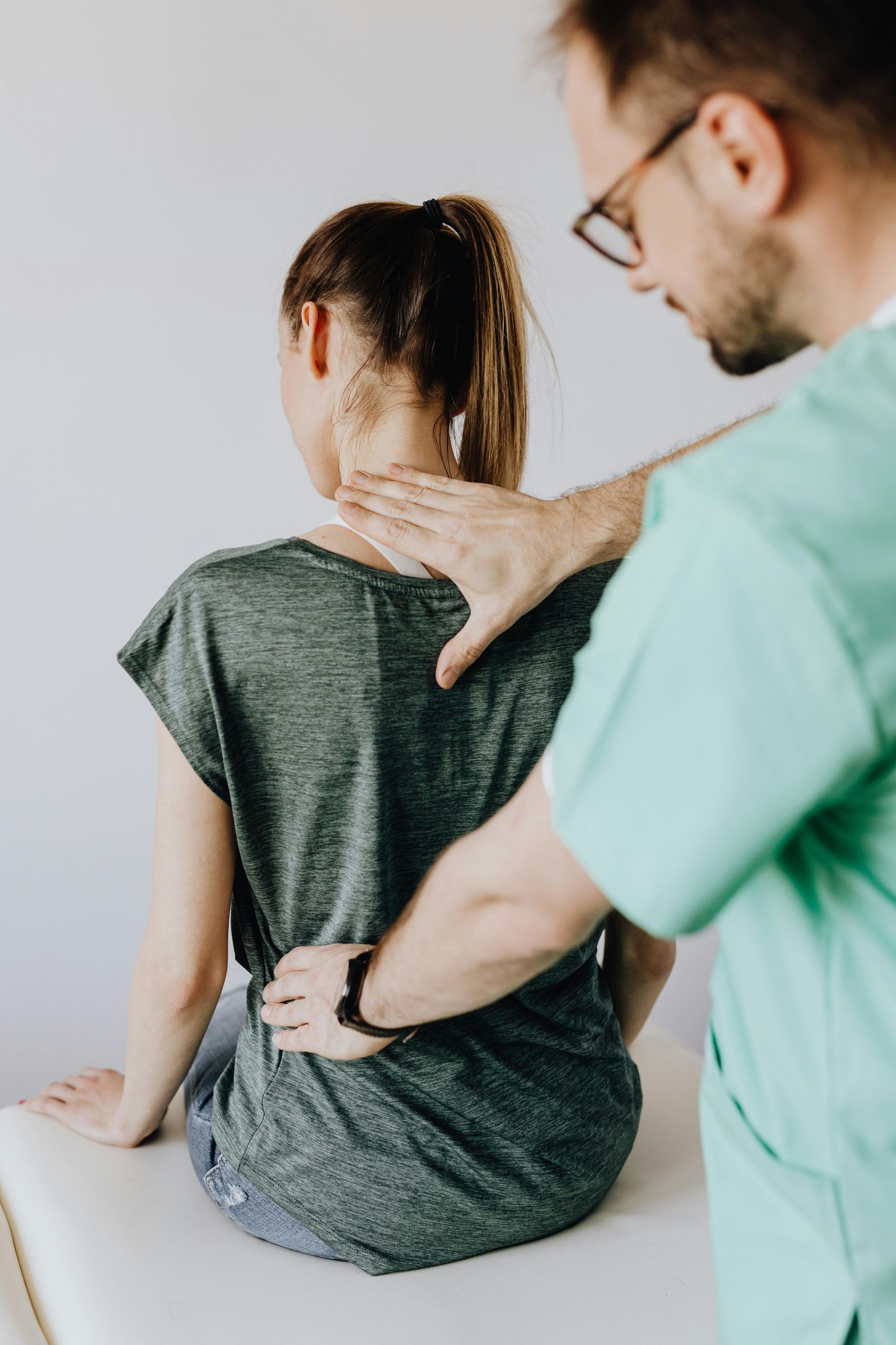 A healthcare professional in green scrubs performing a physical assessment on a patient’s back in a clinic.