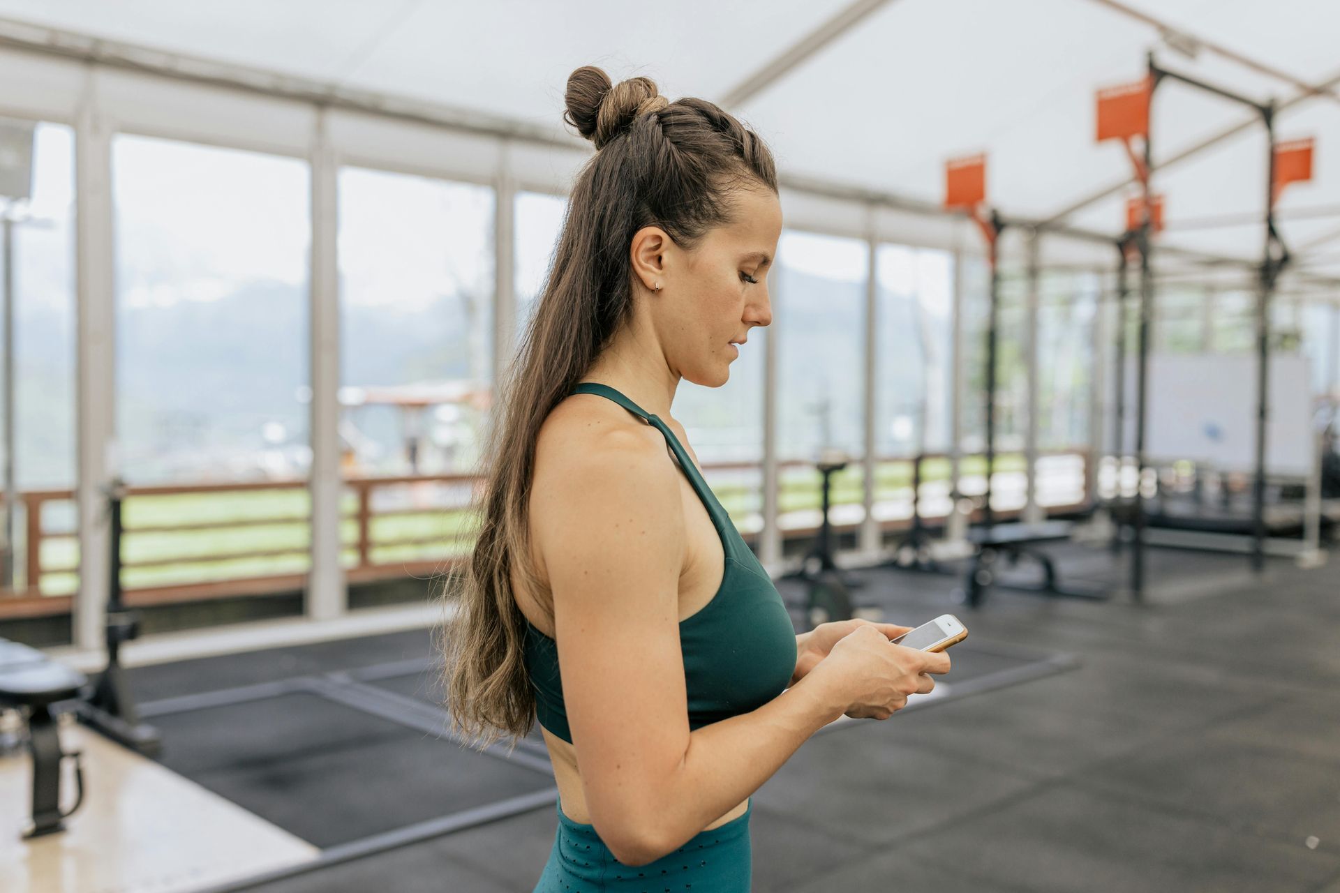 A person wearing a green fitness set uses a smartphone while standing inside a bright, open gym.