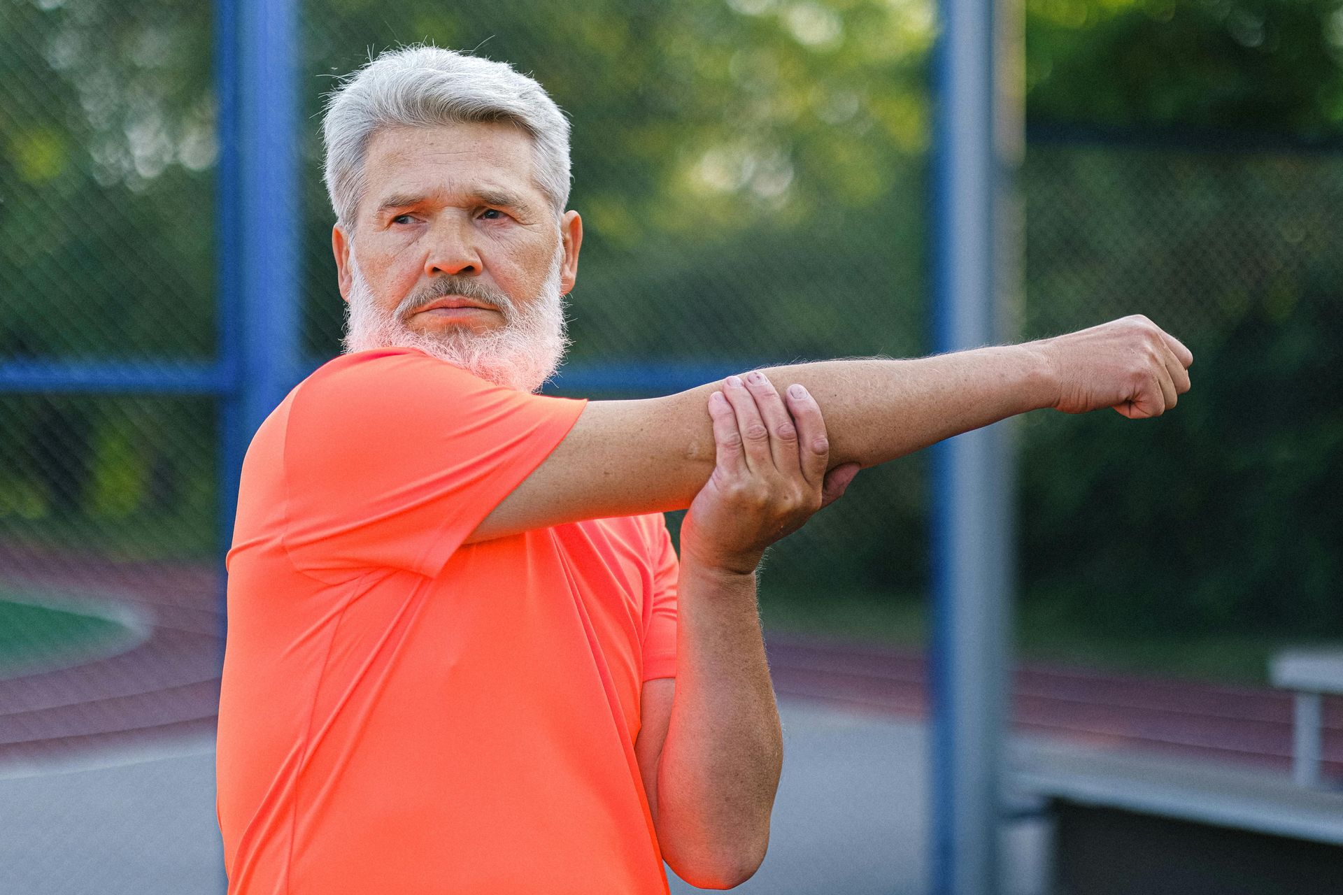 An active person in an orange shirt stretches their arm across their chest outdoors on a sports court.