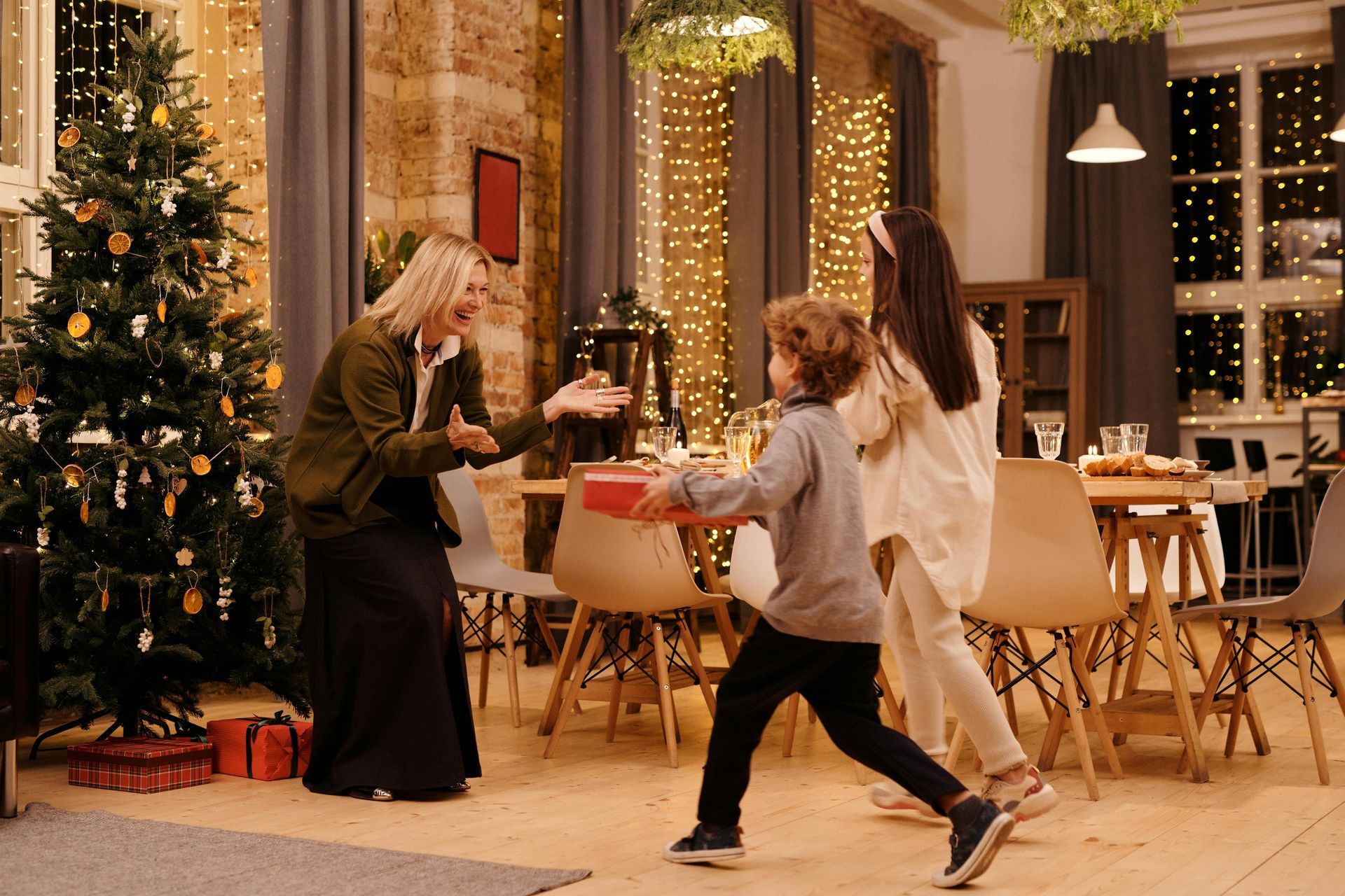 A person happily receives a gift from a child near a lit Christmas tree in a cozy room with dining furniture.