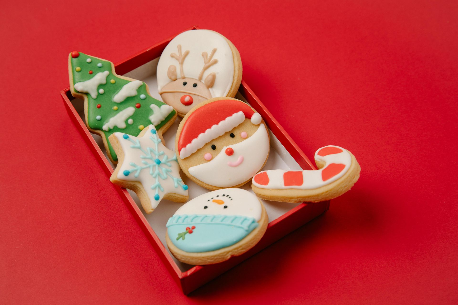A red gift box containing festive Christmas-themed sugar cookies shaped like a tree, star, reindeer, Santa, snowman, and cane.