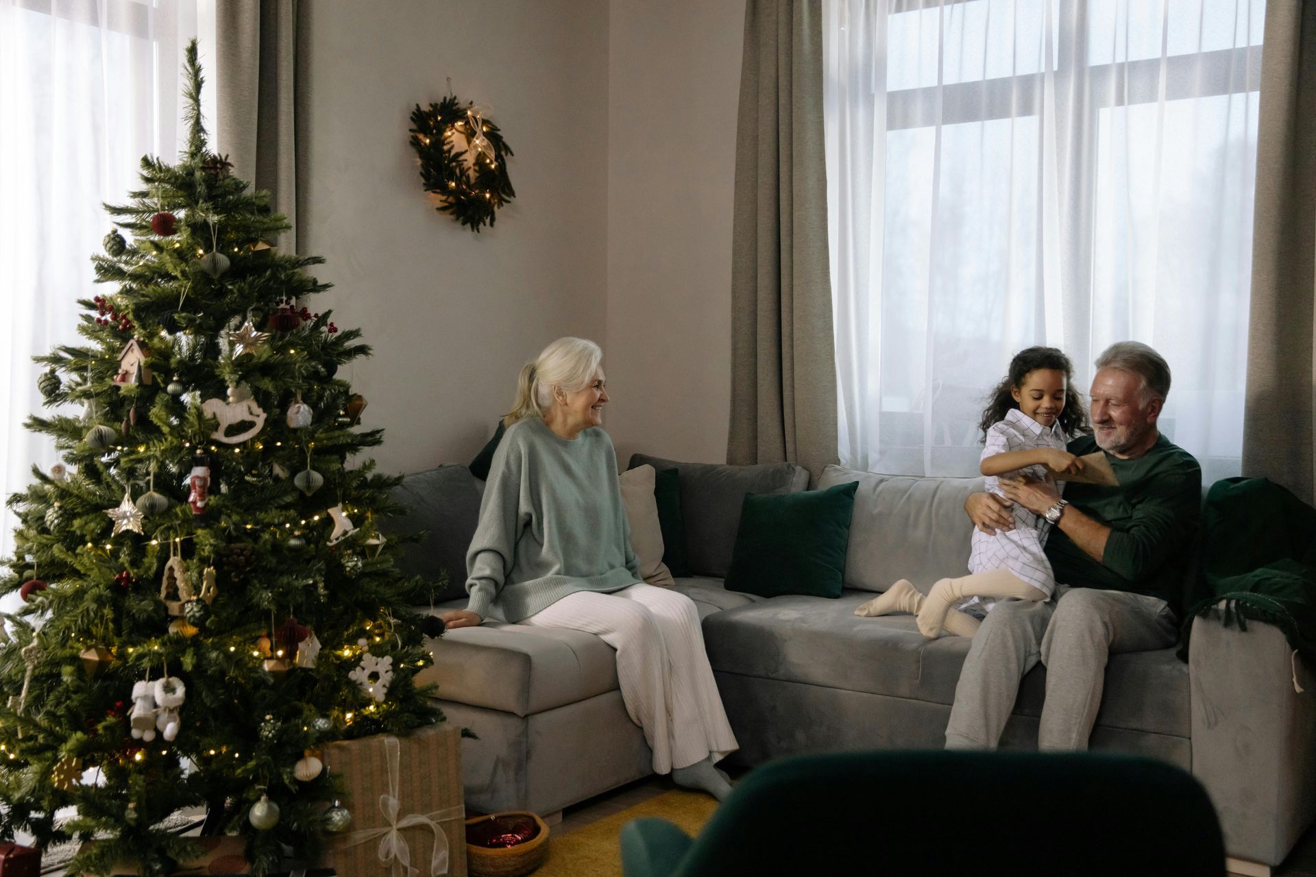 An older couple and a child sit on a couch in a living room decorated with a Christmas tree, sharing a joyful moment.