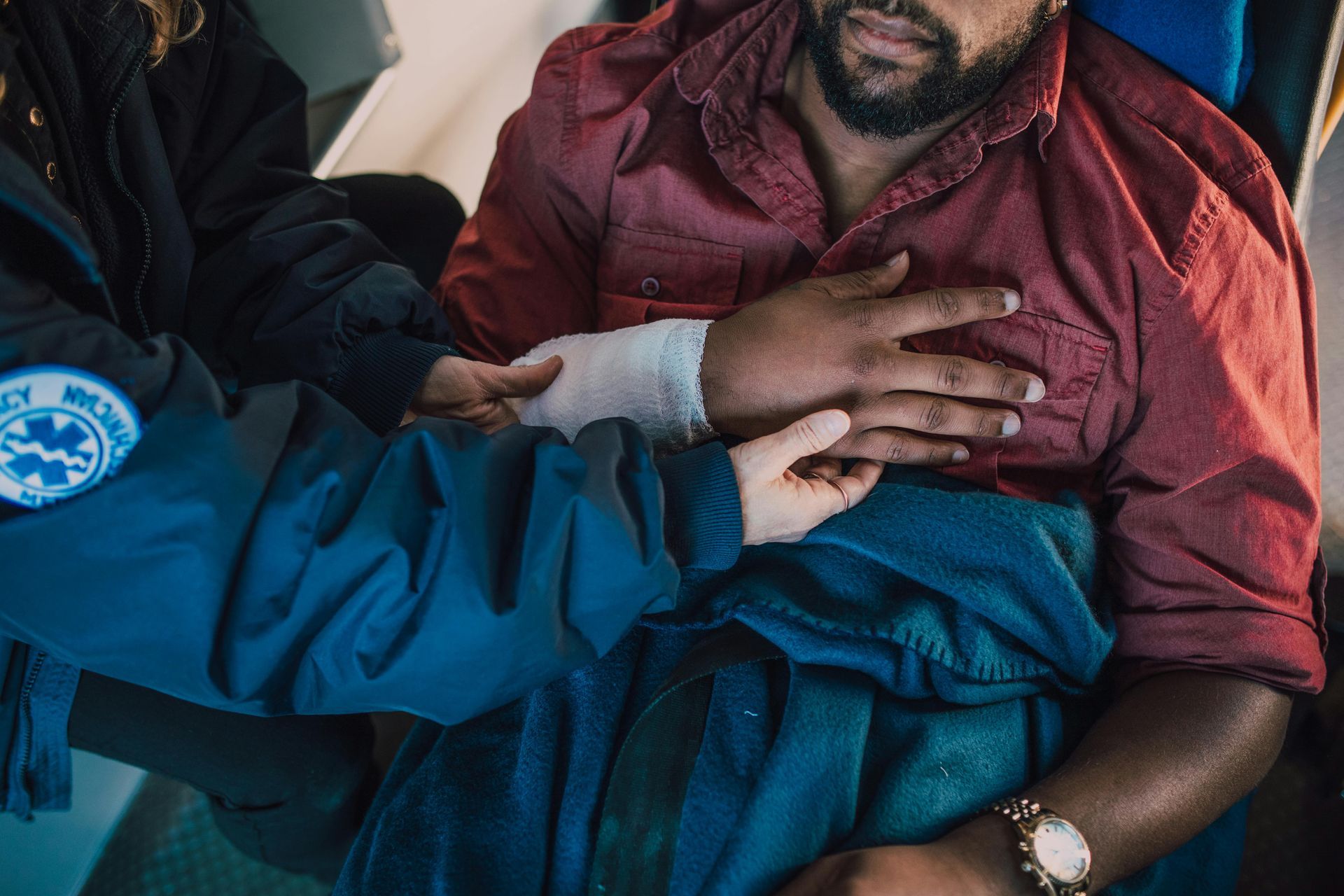 A medic in a blue uniform attends to a person with a bandaged hand, wrapping them in a blanket inside an ambulance.