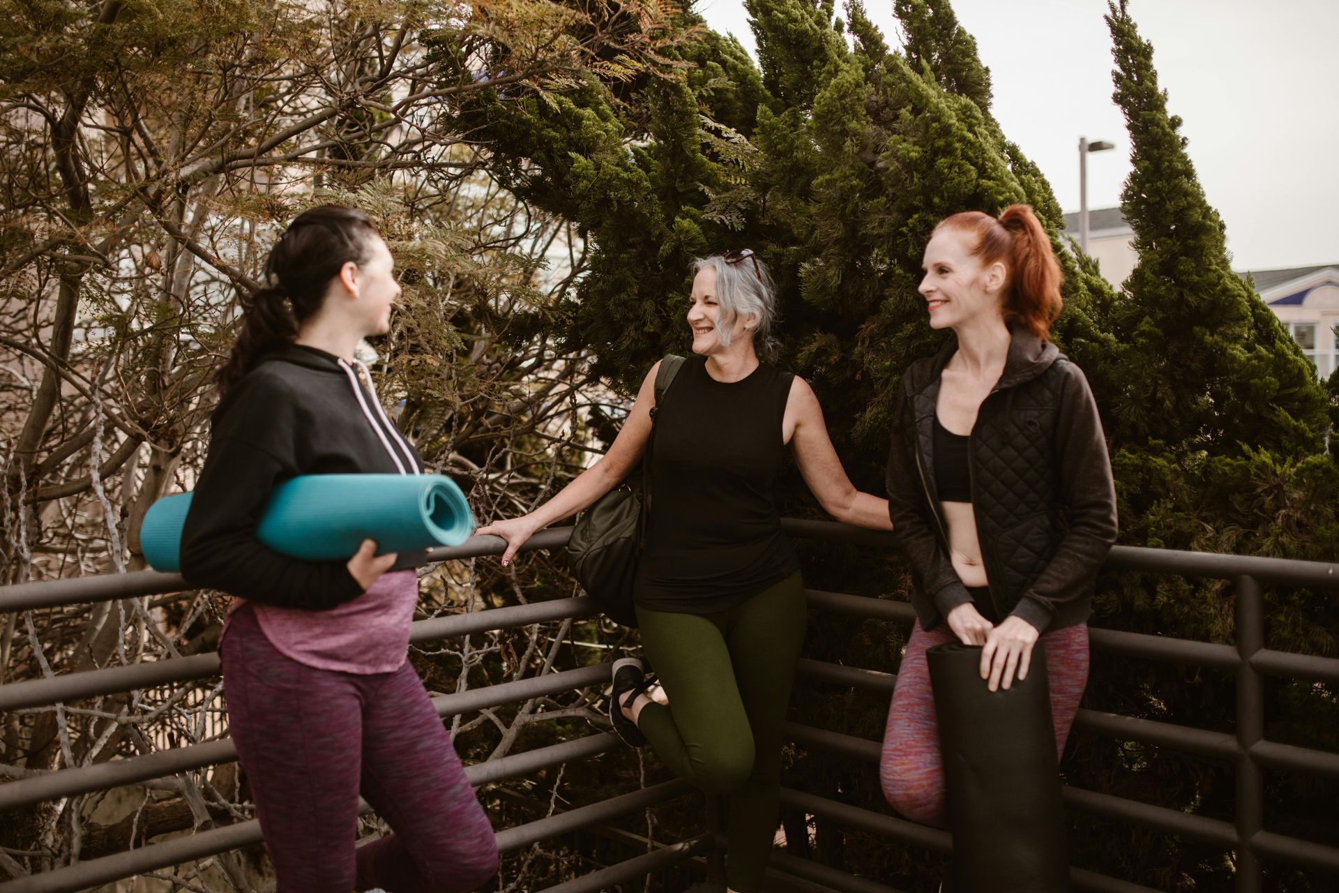 Three people in athletic wear stand by a railing outdoors, talking while holding rolled-up yoga mats.