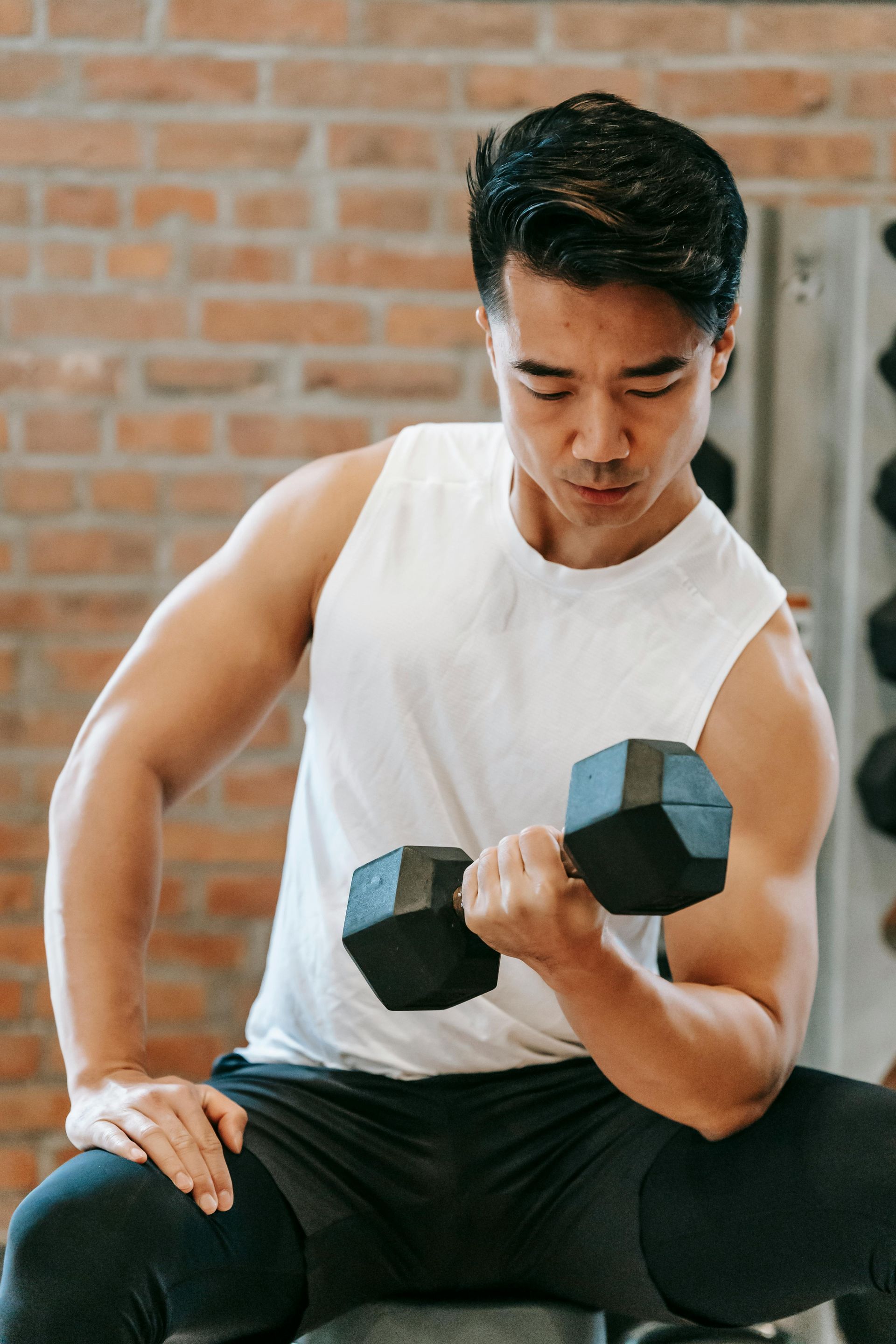 A person wearing a white tank top sits in a gym, focusing while performing a bicep curl with a black dumbbell.