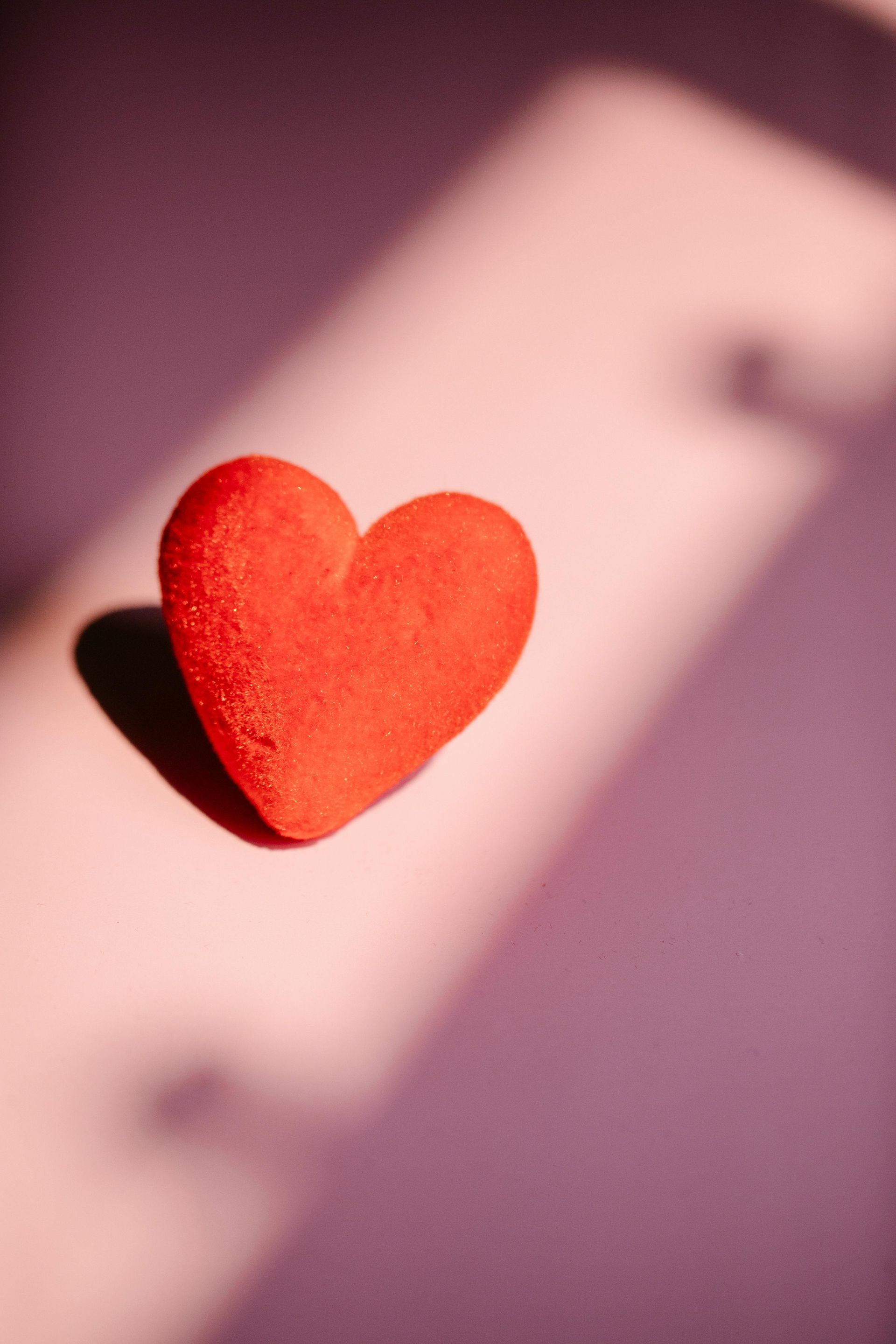 A vibrant red heart-shaped object resting on a light pink surface, illuminated by sunlight with a distinct shadow.