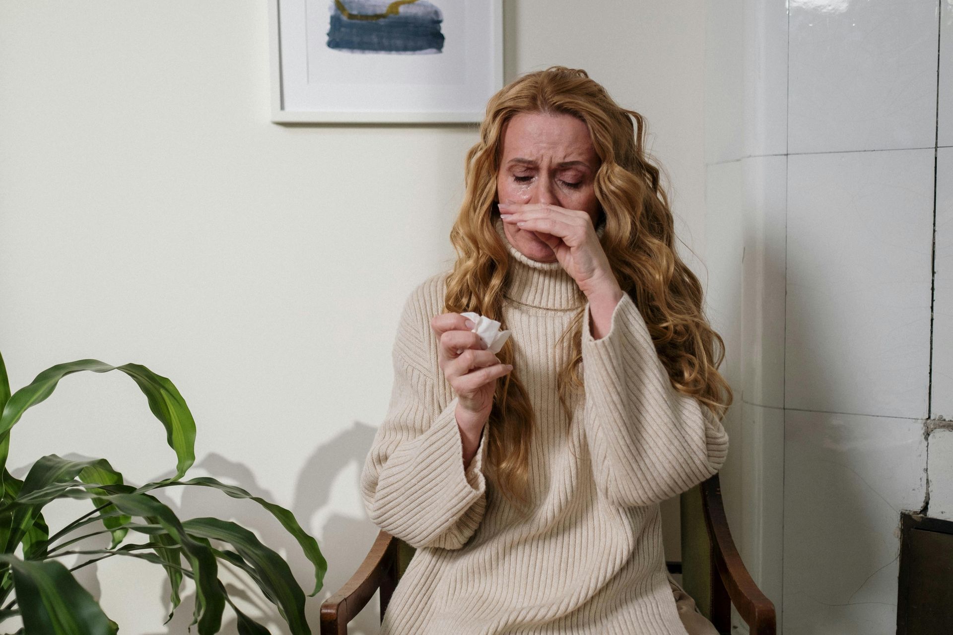 A person with long wavy hair sits in a chair, holding a tissue to their nose, appearing unwell in a room with a plant.