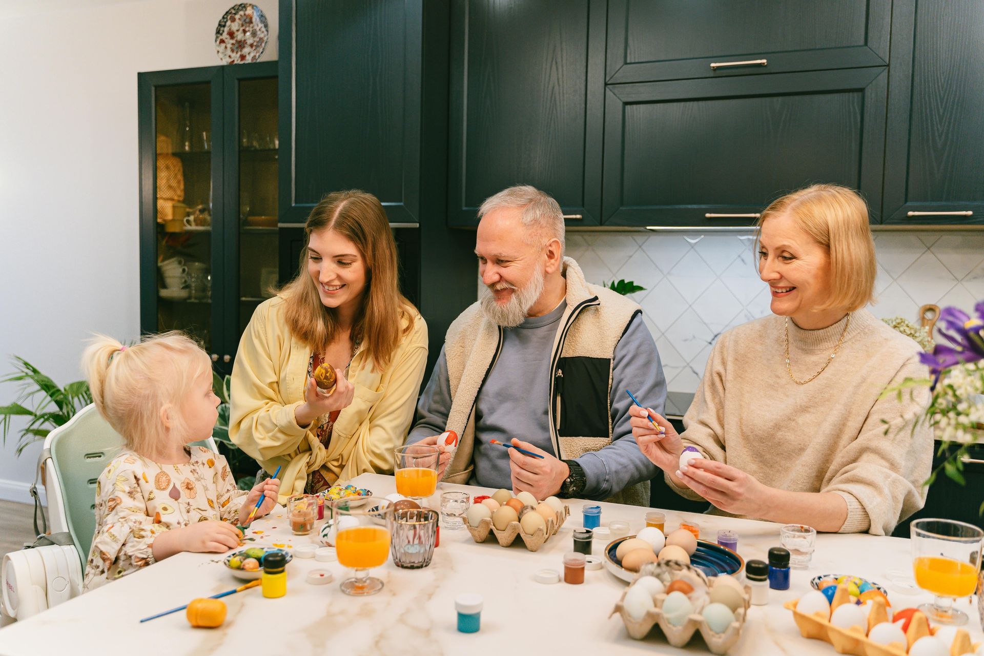 A family sits at a kitchen table painting Easter eggs, with colorful dyes, brushes, and egg cartons spread before them.
