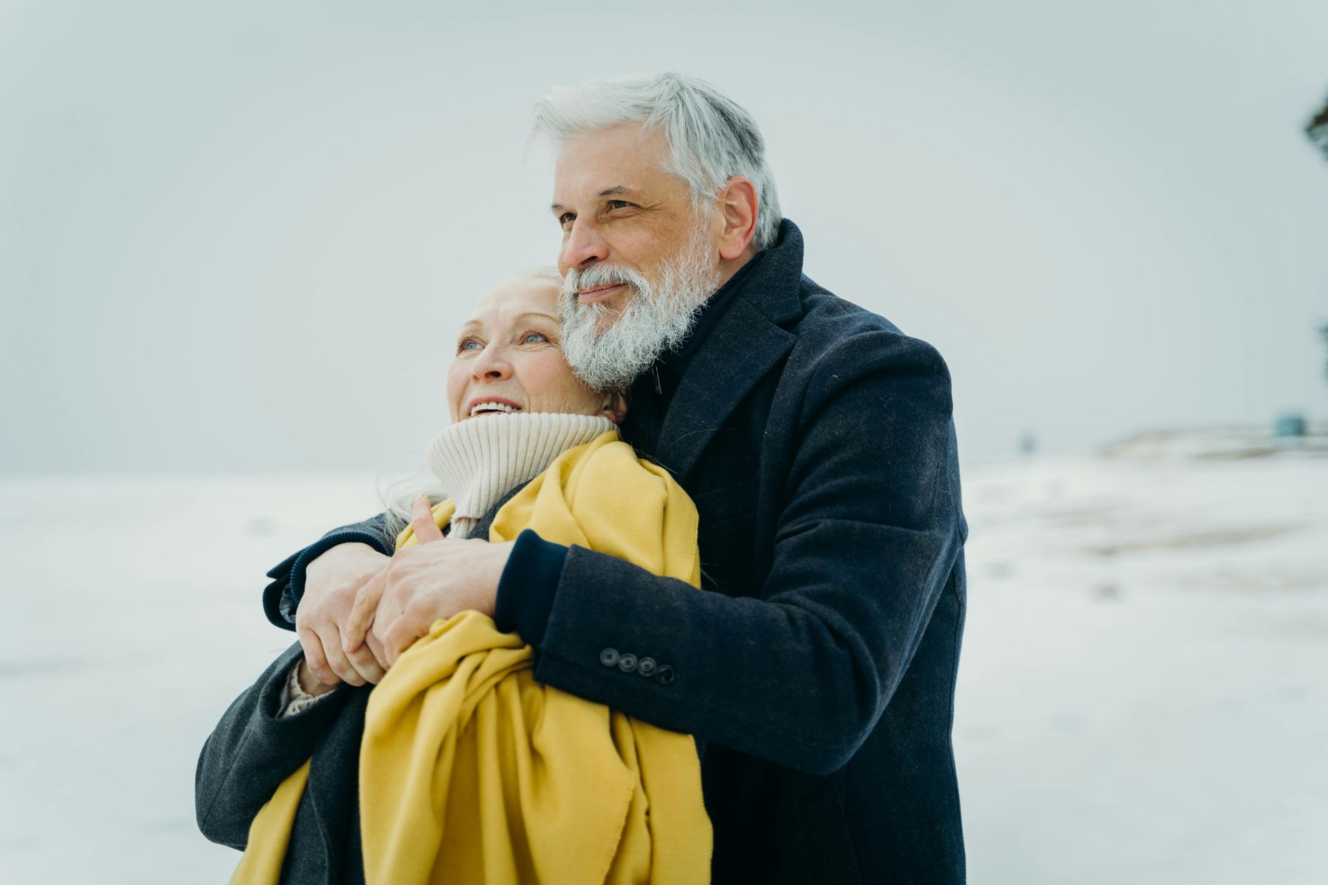 An older couple embraces in a snowy landscape, the person in front wearing a yellow blanket and the other in a dark coat.