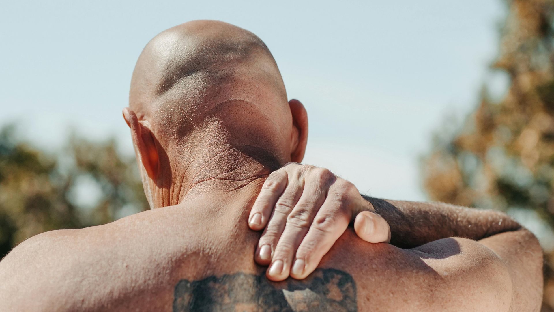A person viewed from behind, reaching up with their right hand to touch the back of their neck and upper shoulder area.