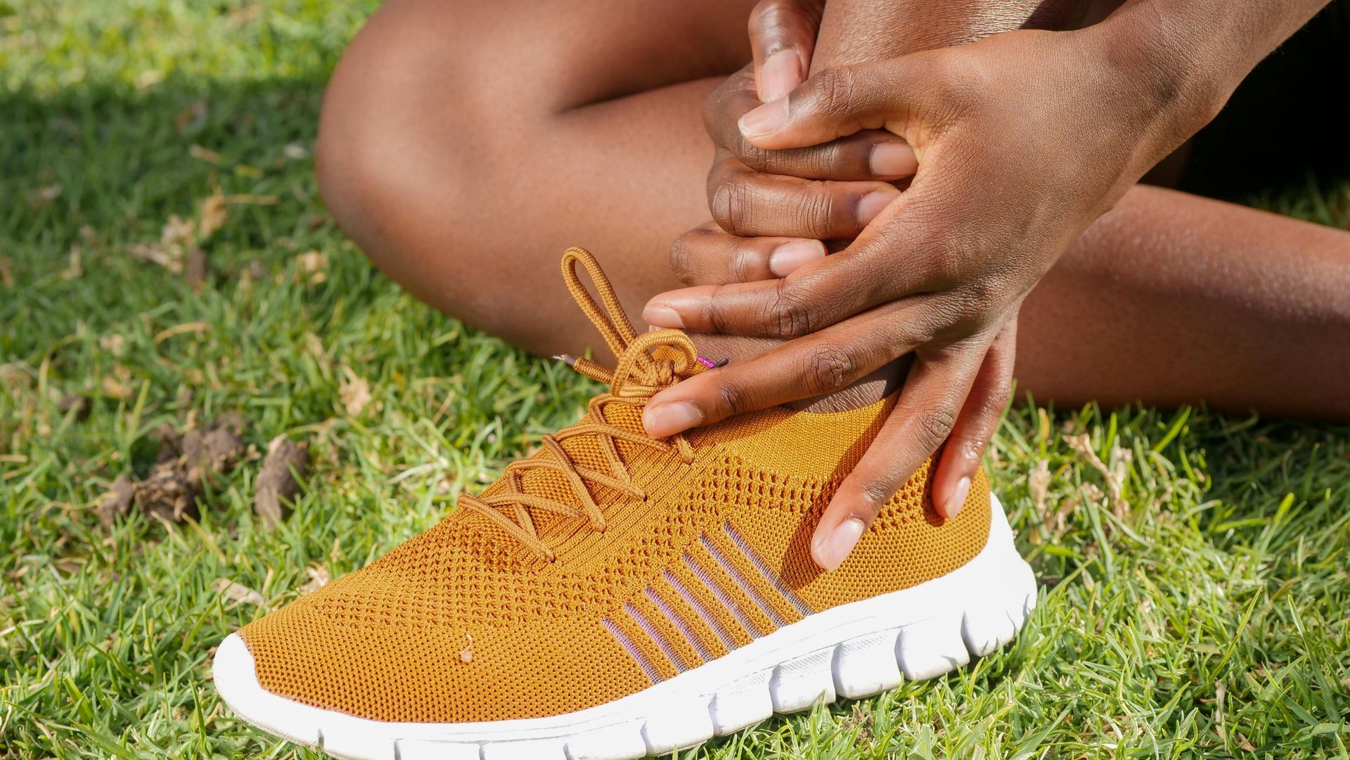 A person sitting on grass, holding their ankle with both hands, suggesting pain or injury.