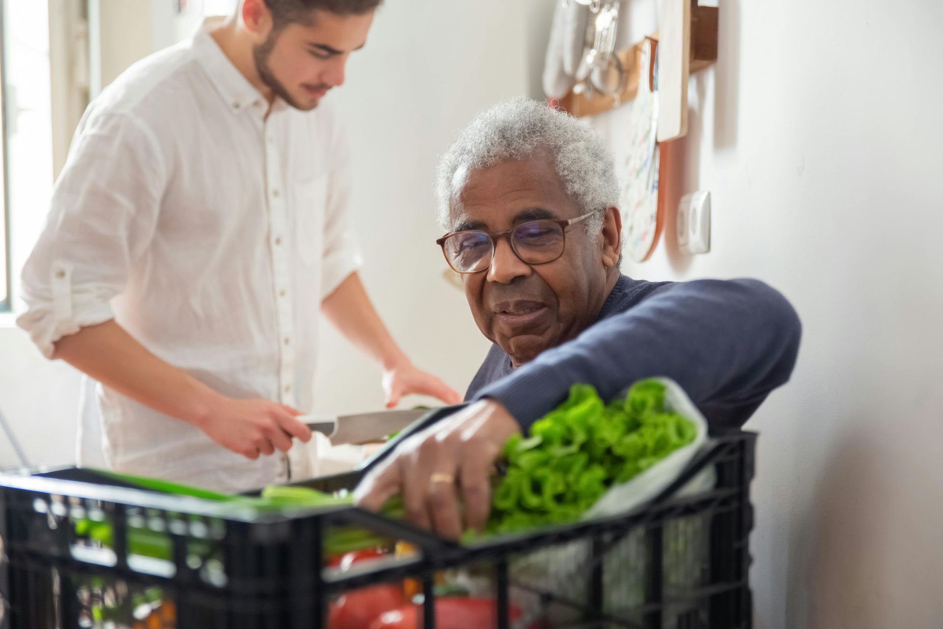 A person reaches into a black crate of fresh produce while another person uses a knife in the background.