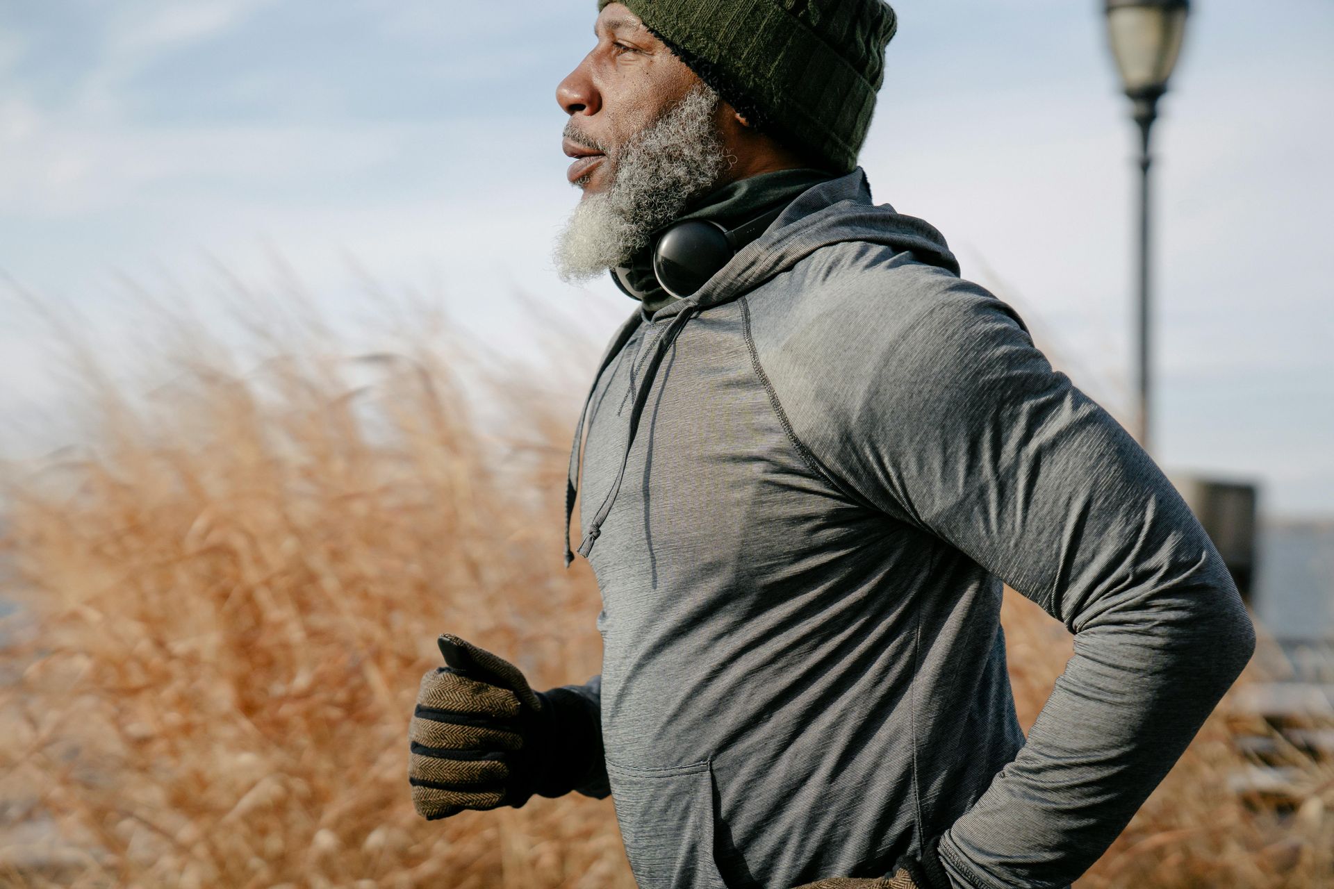 A person with a grey beard, wearing a green beanie and grey hoodie, runs outdoors beside tall, dry grass.
