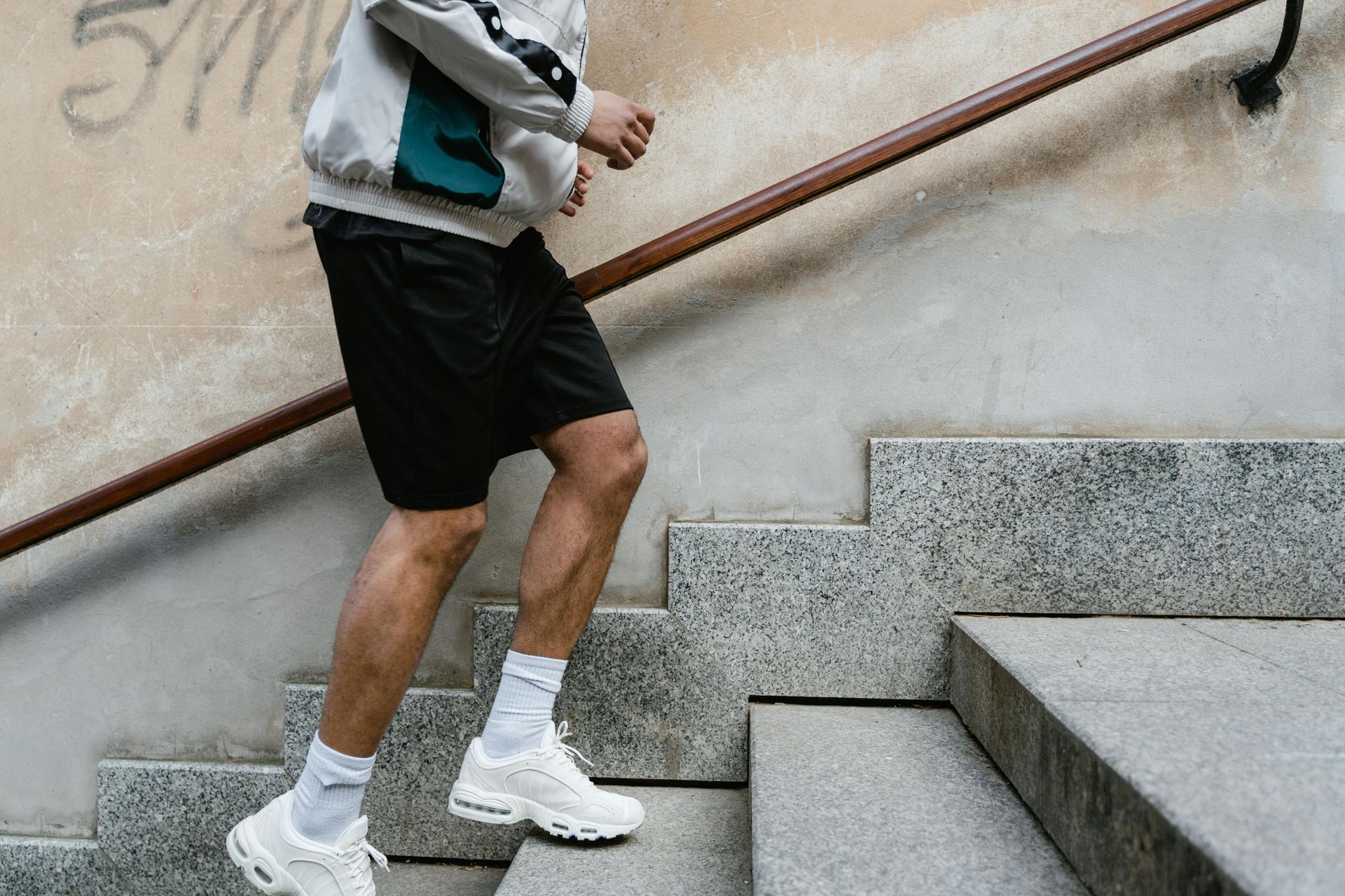 Person in a light jacket and black shorts walking up outdoor stone stairs holding a handrail.