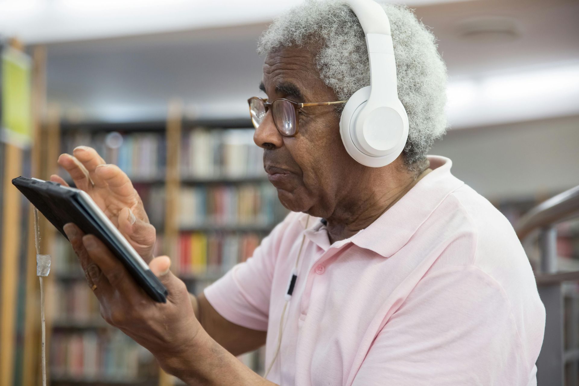 An individual wearing white over-ear headphones uses a tablet while standing in a library.
