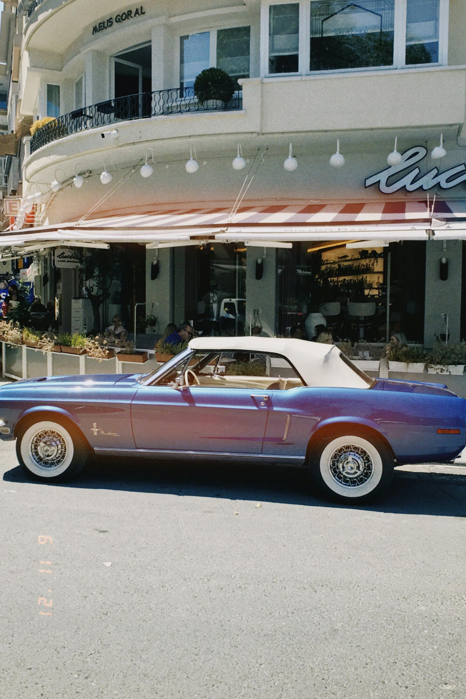 A vintage blue convertible with a white top parked in front of a building with a striped awning.