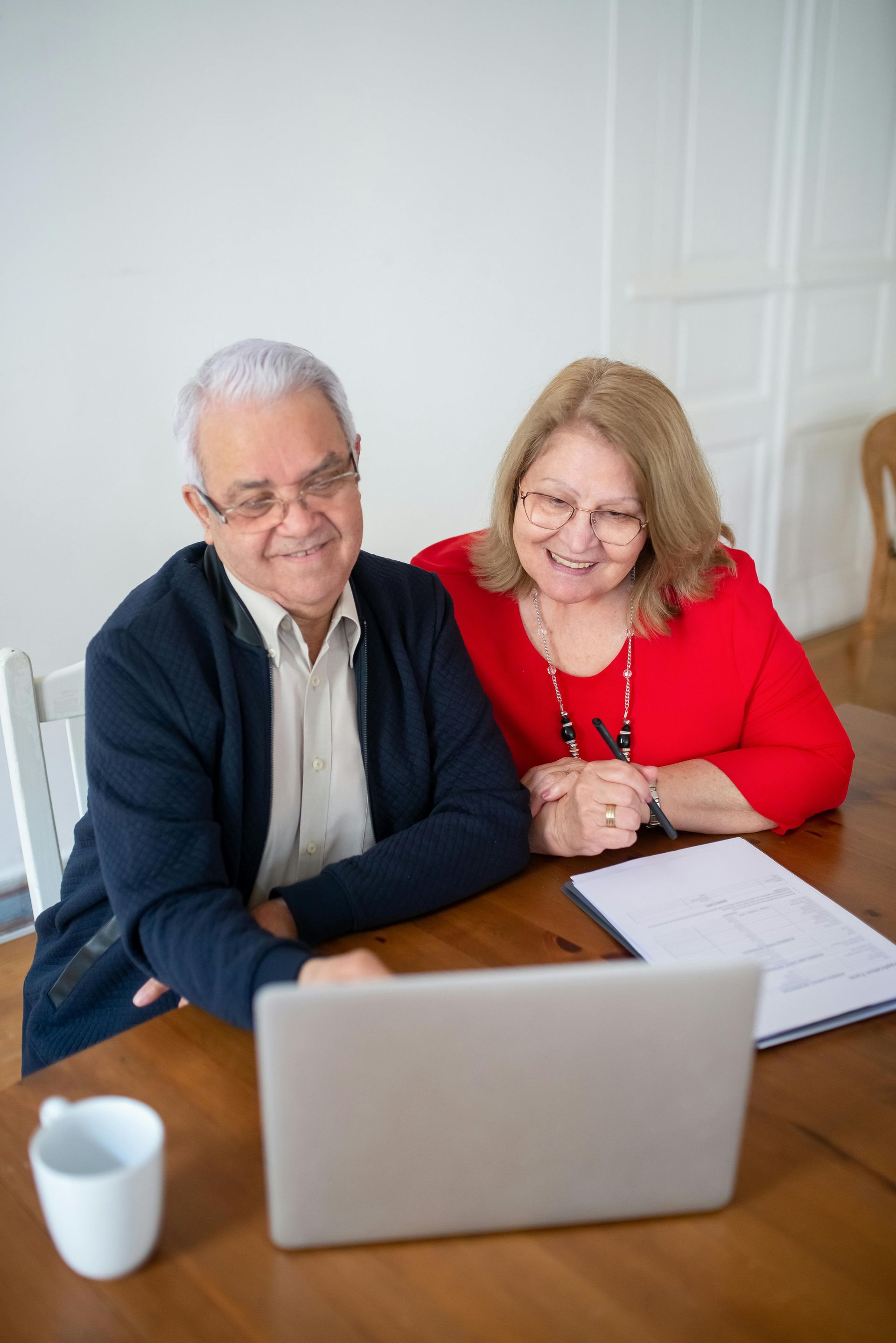 Two people sitting at a wooden table looking at a laptop together, with a white mug and papers nearby in a bright room.