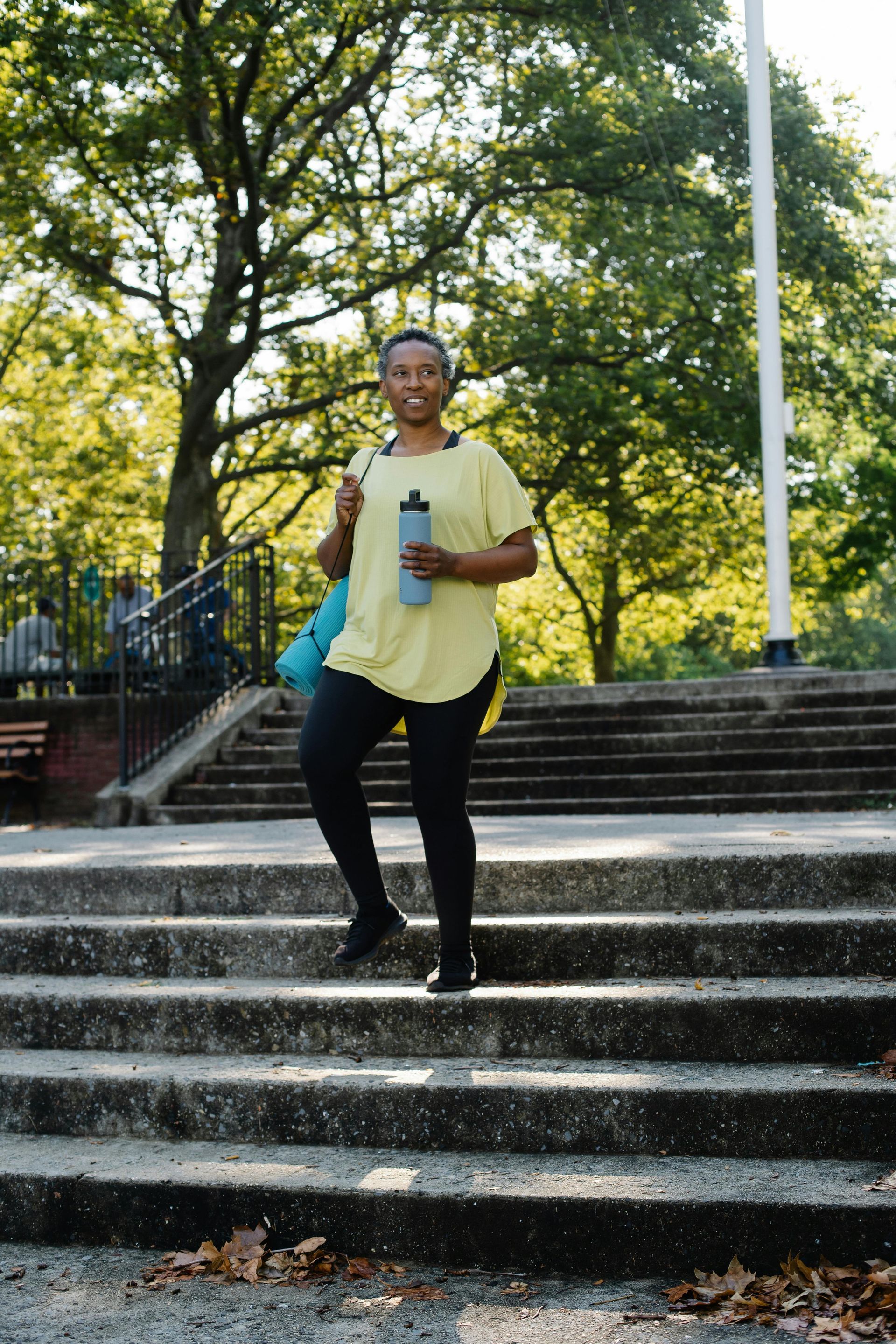 A person in a yellow shirt and black leggings walks down stone stairs in a park, holding a water bottle and yoga mat.