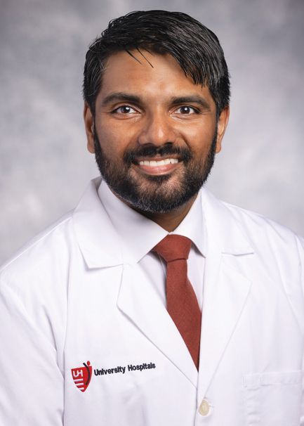A smiling person in a white lab coat with a University Hospitals logo and a red tie, posed against a gray backdrop.