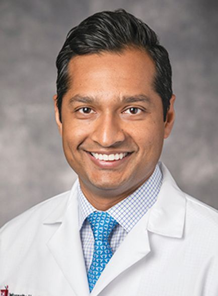 A smiling professional in a white doctor’s coat, patterned dress shirt, and tie against a mottled grey background.