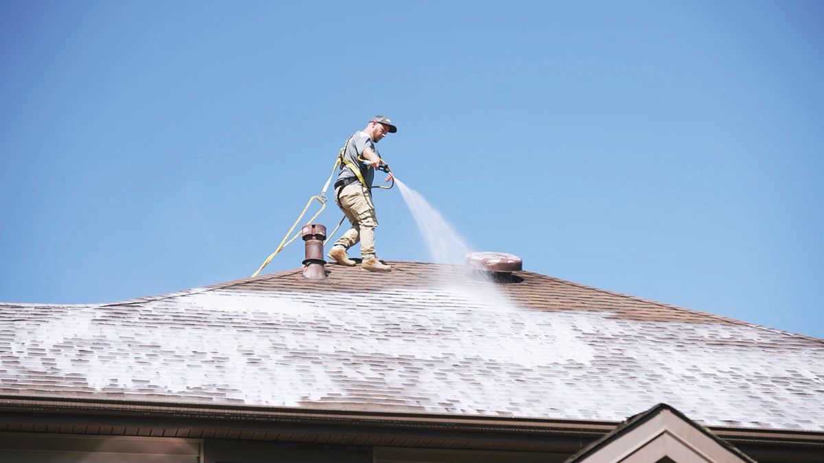A person on a sloped roof uses a pressure washer to spray cleaning solution onto shingles against a clear blue sky.