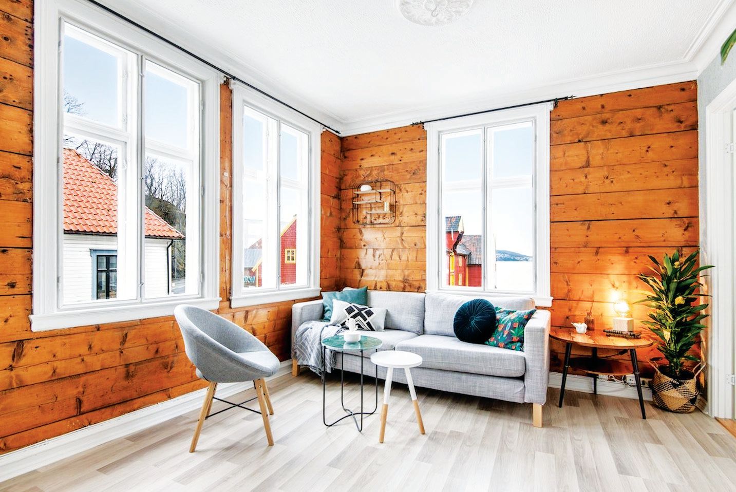 A cozy living room with natural wood walls, a light gray sofa, a mid-century modern chair, and windows with outdoor views.