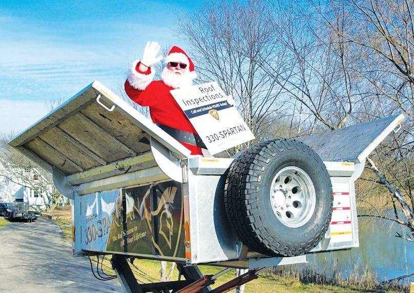 Santa Claus waves from a parked dump trailer while holding a Roof Inspections sign against a background of trees and a pond.