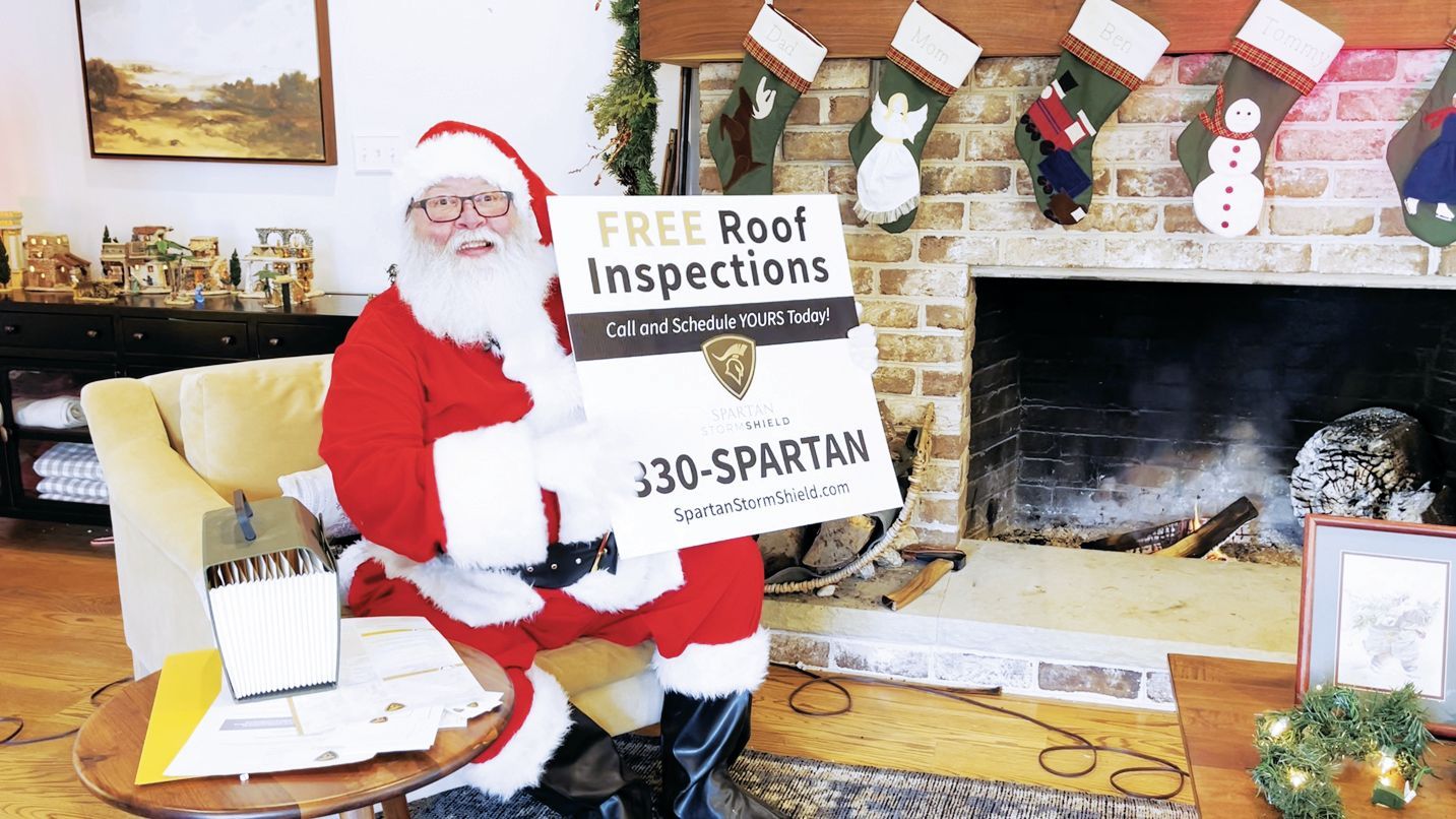Santa Claus sits in a living room, holding a sign for free roof inspections and the phone number 830-SPARTAN.