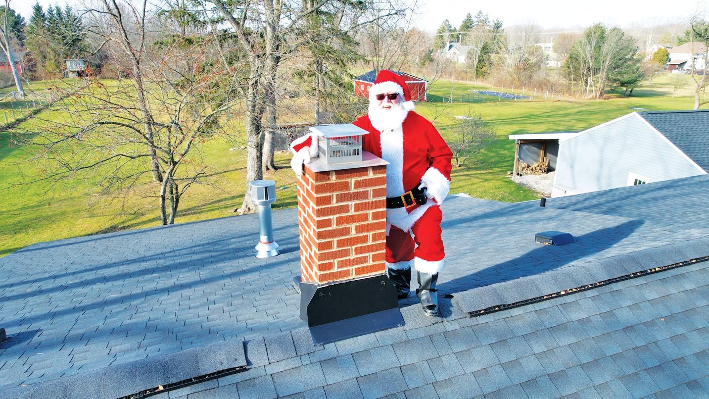 A person dressed as Santa Claus stands on a shingled residential roof next to a brick chimney on a sunny day.