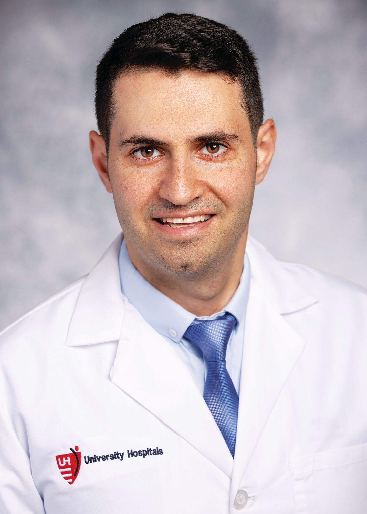 A smiling professional wearing a white doctor's coat and blue tie, positioned in front of a neutral gray background.