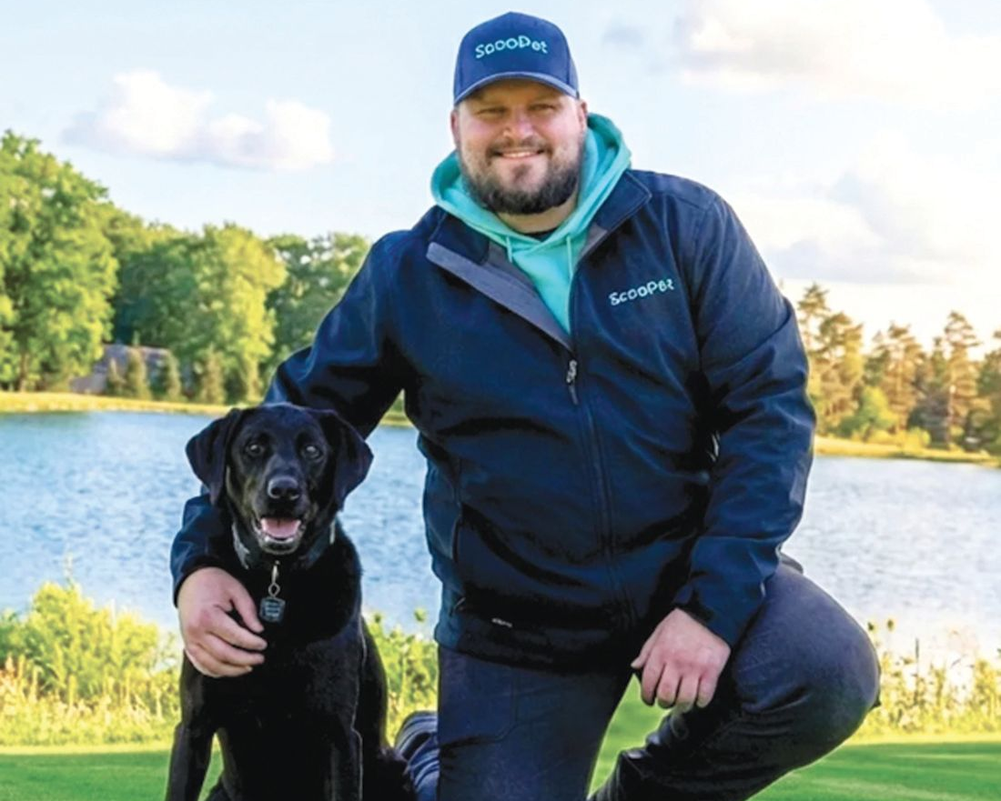 A smiling person in a blue jacket and cap kneels in a grassy field by a lake, with their arm around a black dog.
