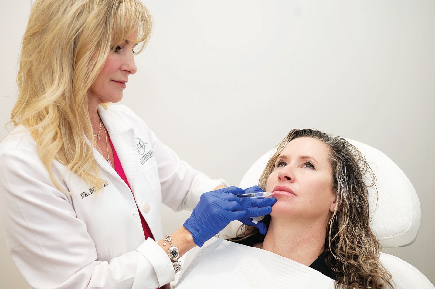 A medical professional in a white coat and blue gloves performs a cosmetic injection on a patient's lip in a clinic.