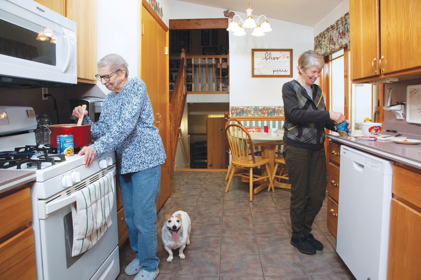 Two people prepare food in a kitchen while a small dog stands between them.