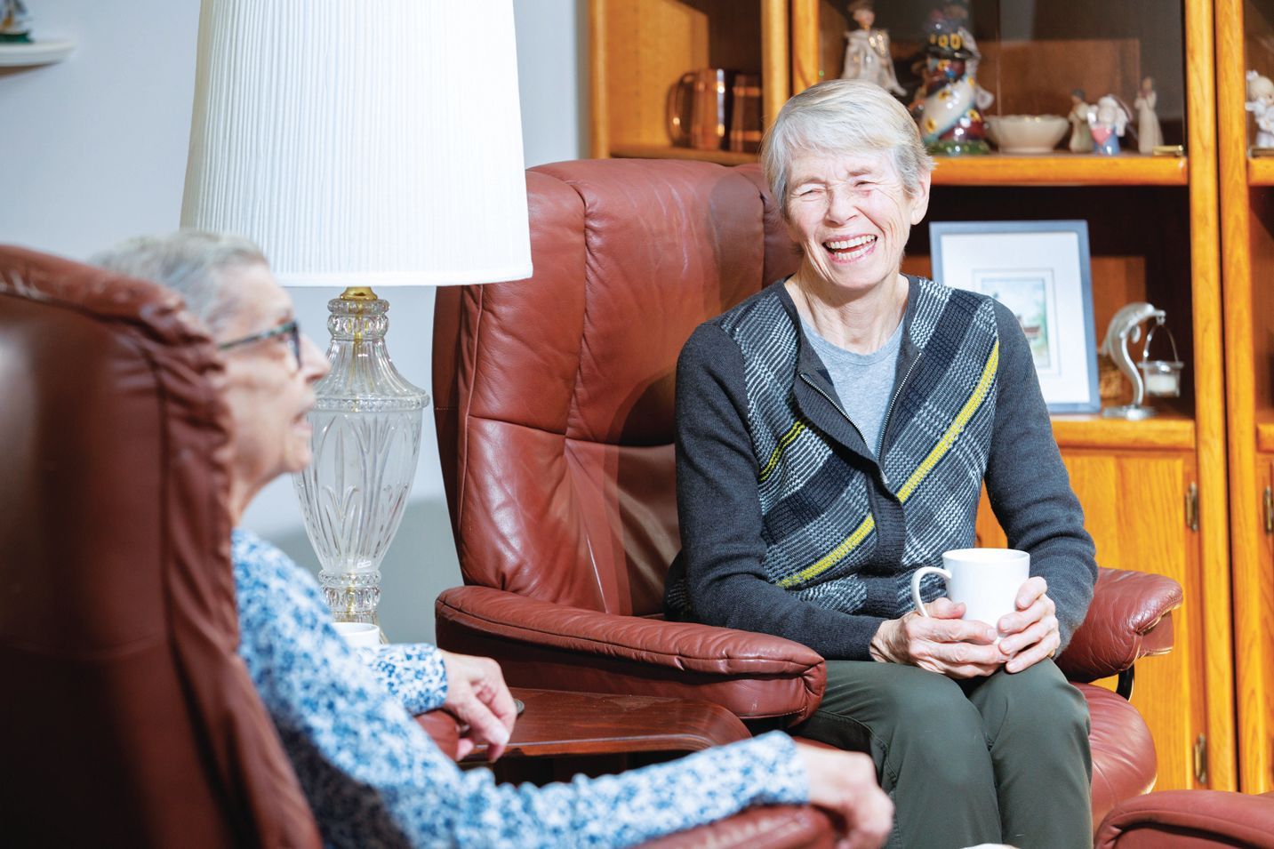 Two people sit in leather chairs in a living room, talking and laughing while holding a mug.