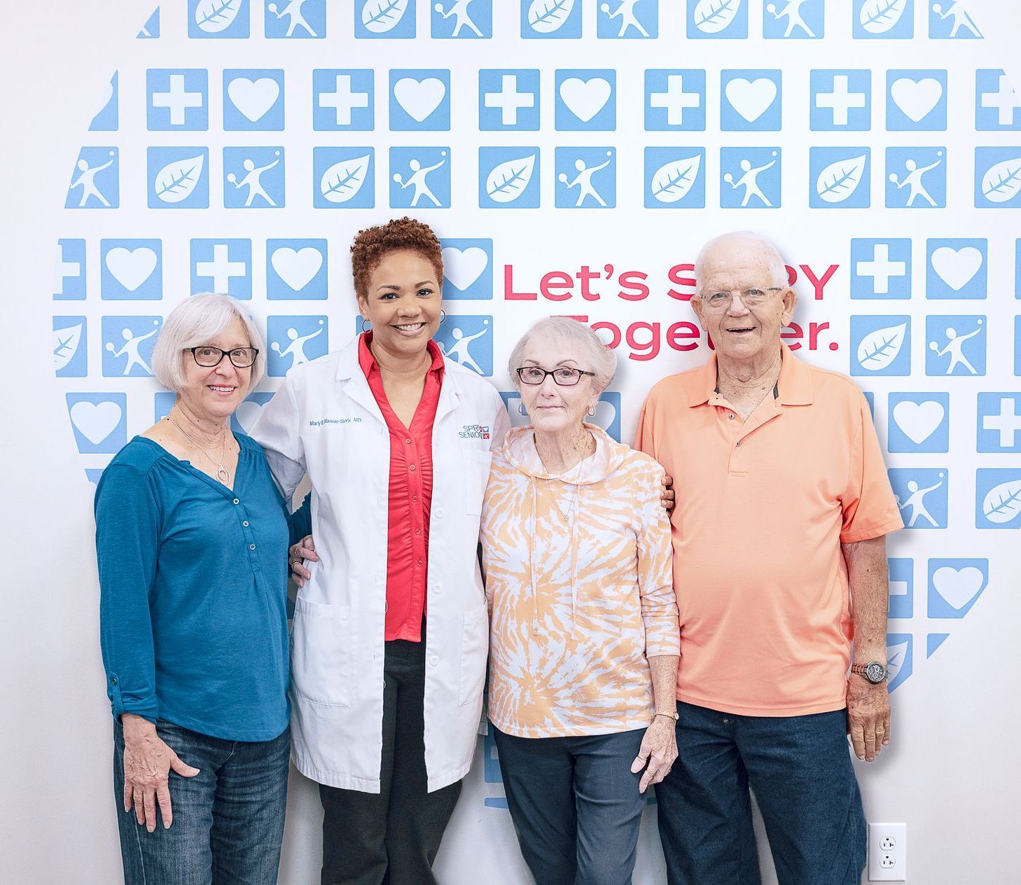 A healthcare professional in a white coat stands between three smiling adults in front of a blue and white patterned wall.