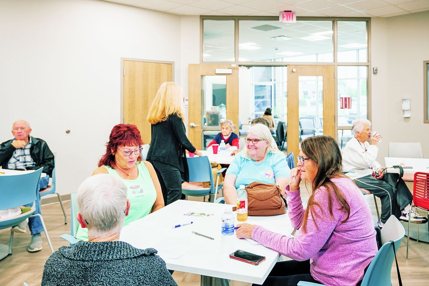 A group of people seated at tables in a brightly lit community room, engaged in conversation during an informal gathering.