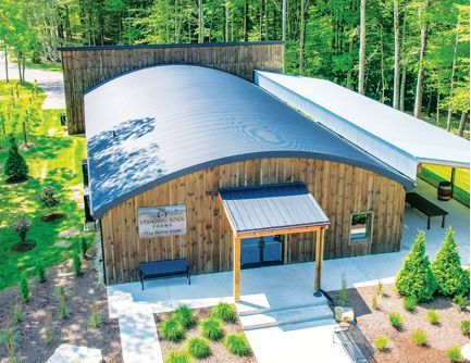 An aerial view of a rustic, wood-sided building with a curved metal roof and a small covered entrance, set in a forest.