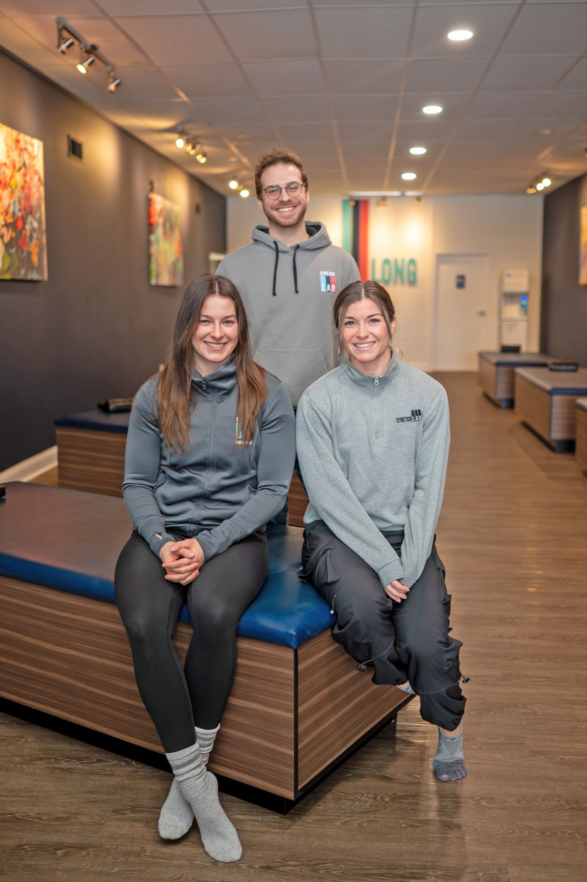 Three people smiling in a bright, modern studio space; two sit on a padded bench while one stands behind them.