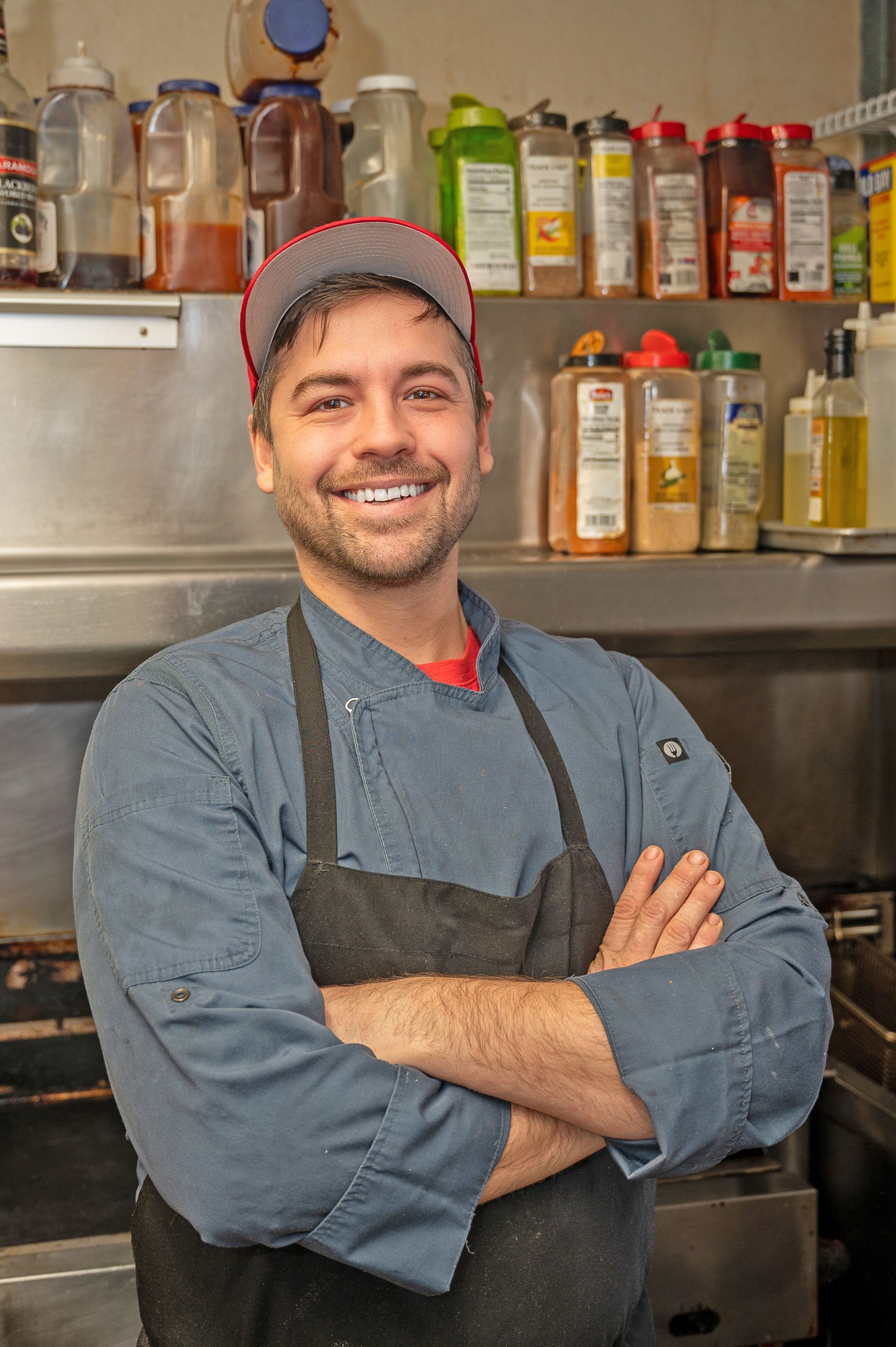 A smiling chef in a blue coat and apron stands with arms crossed in a kitchen, with spice containers on shelves behind.