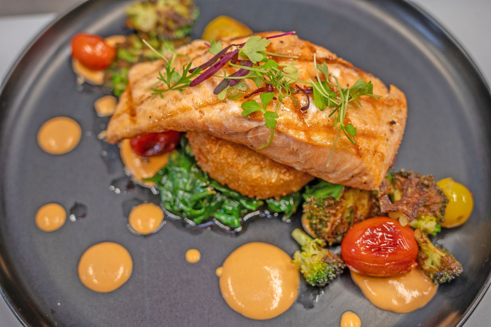 A grilled salmon fillet sits on a crispy patty and sautéed spinach, served with roasted broccoli, tomatoes, and sauce.