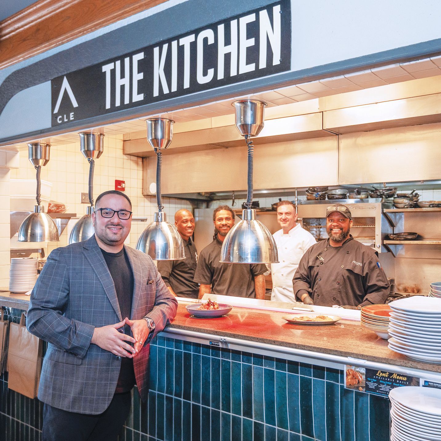 A man in a blazer stands in front of a restaurant kitchen counter with a team of cooks smiling behind him.