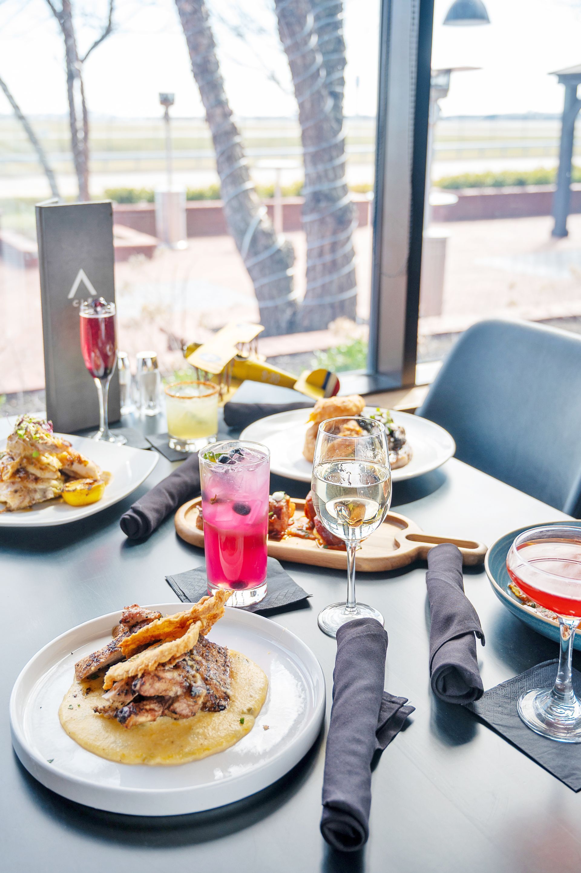 A spread of appetizers, entrees, and colorful cocktails on a restaurant table overlooking a sunny patio.