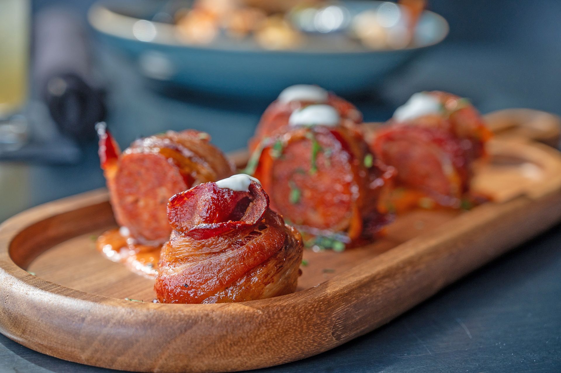 Four bacon-wrapped sausage bites with a small dollop of cream, served on an oval wooden platter.