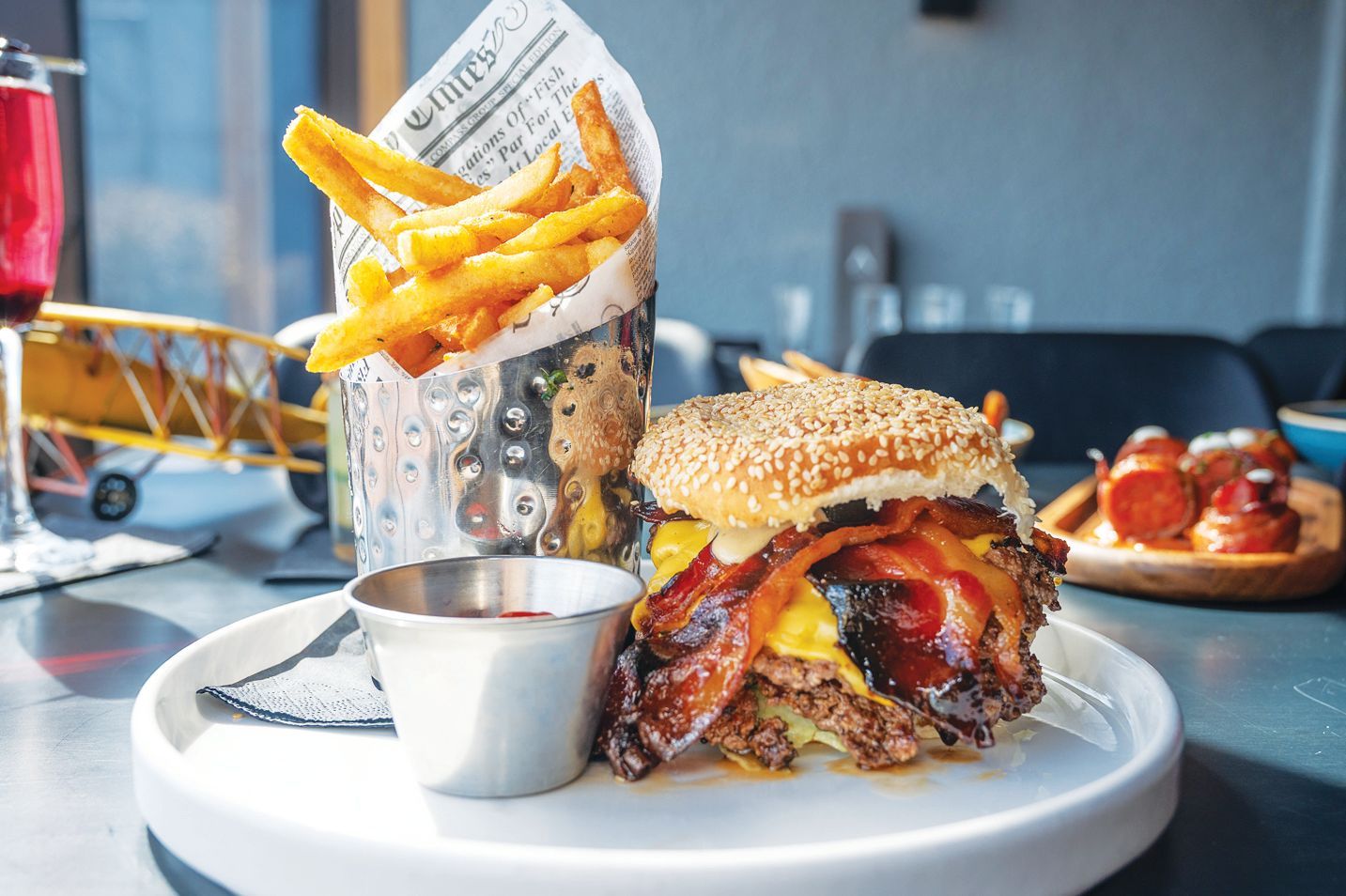 A sesame seed burger topped with bacon and cheese, served with fries in a metal cup and a small side of sauce on a plate.
