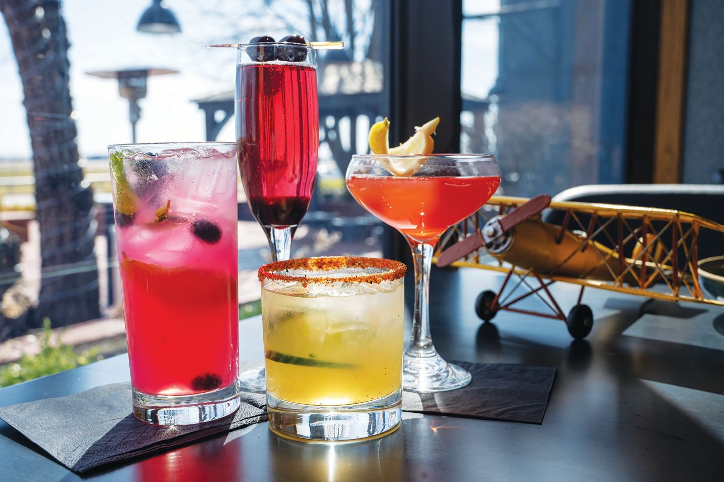 Four colorful cocktails, including a tall pink drink, a champagne flute, a rocks glass, and a coupe, on a table near a window.