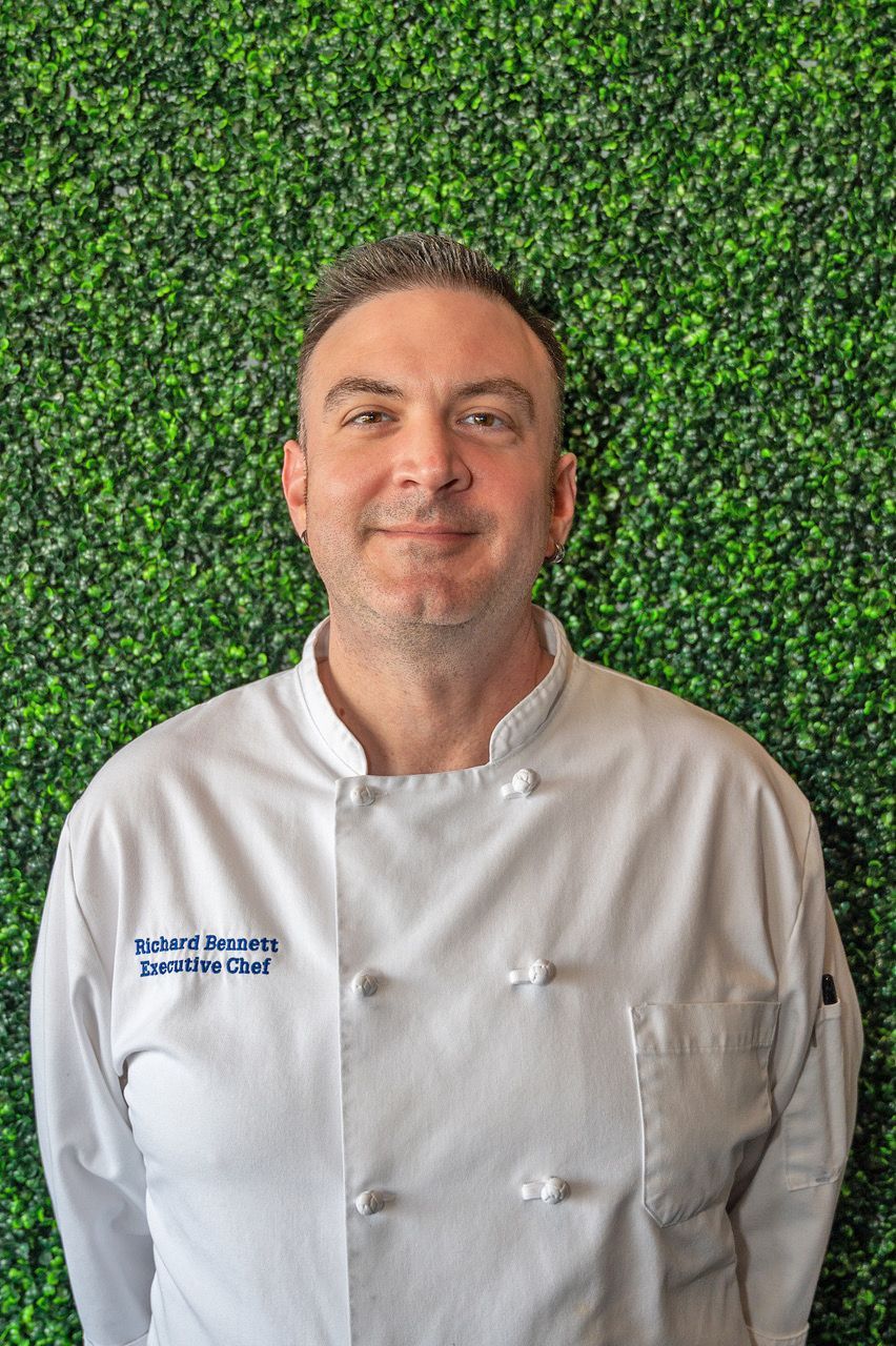 A smiling chef in a white uniform stands in front of a green artificial grass wall.