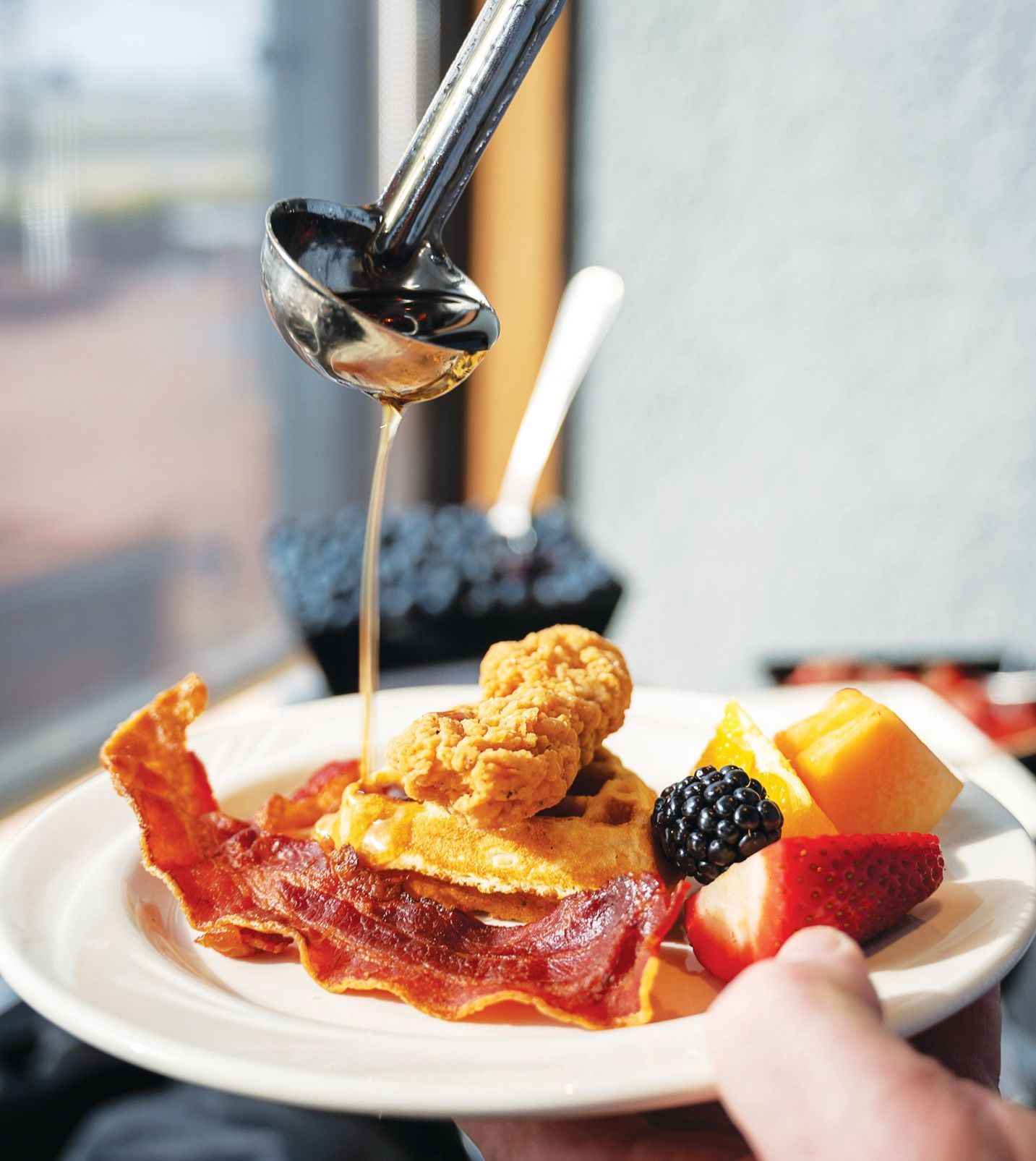 Syrup pours from a ladle over chicken and waffles, served on a white plate with bacon, blackberry, and fruit slices.
